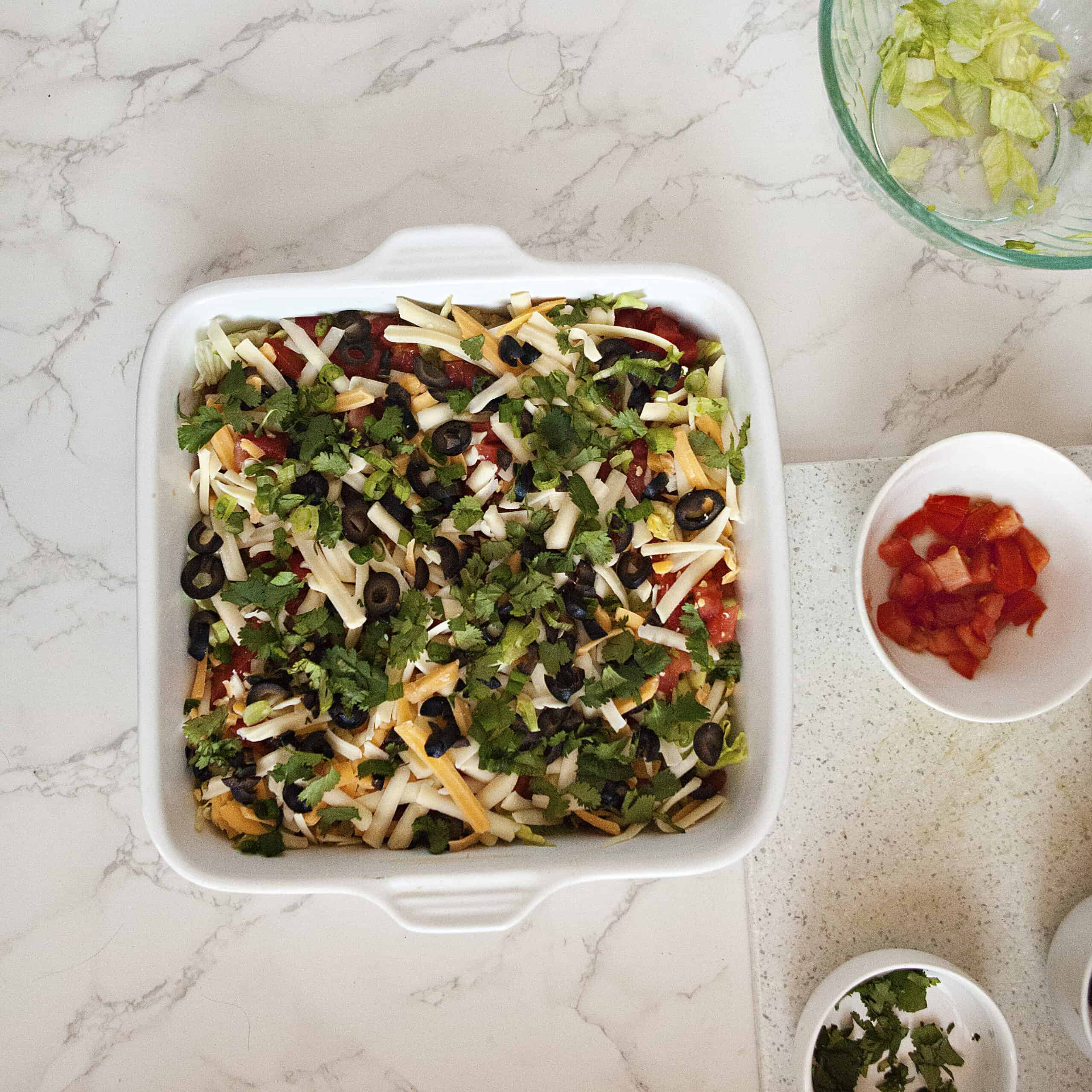 A white square baking dish containing cream cheese taco dip alongside more toppings in small bowls, on a white marble background.