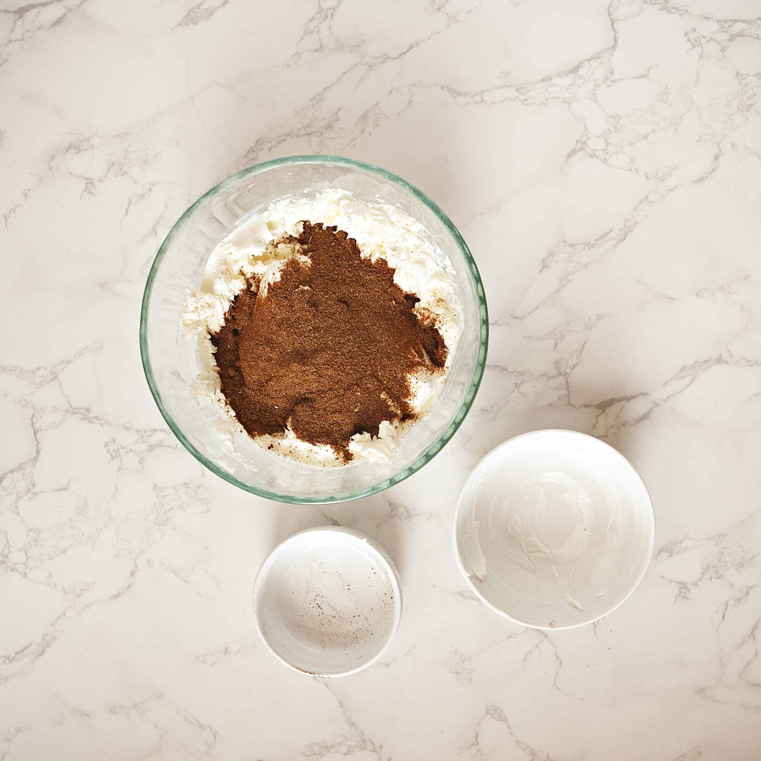 Cream cheese, sour cream, and taco seasoning in a glass bowl next to 2 empty smaller bowls on a white marble background.