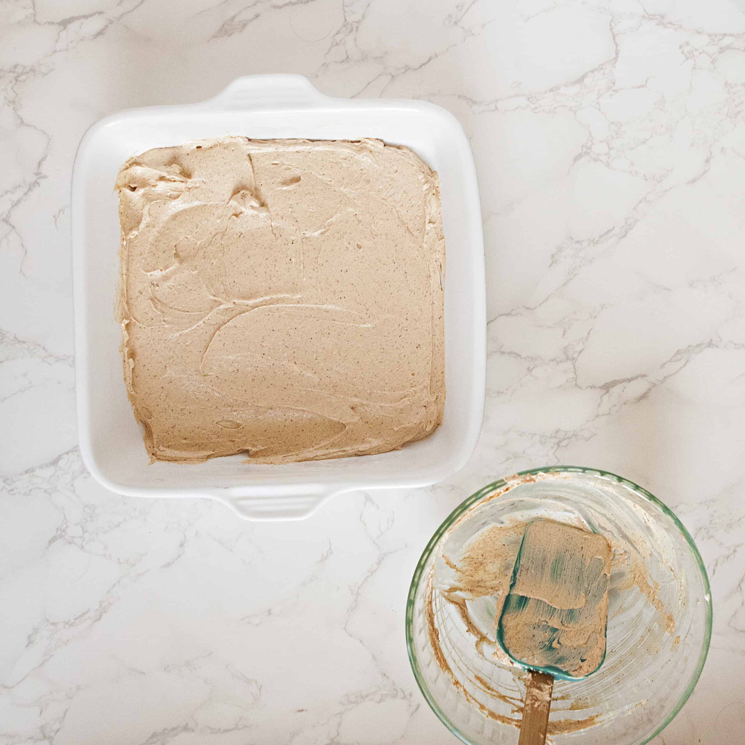 A 9x9 inch white baking dish containing a sour cream/cream cheese mix with taco seasoning, next to an empty bowl, on a white marble background.