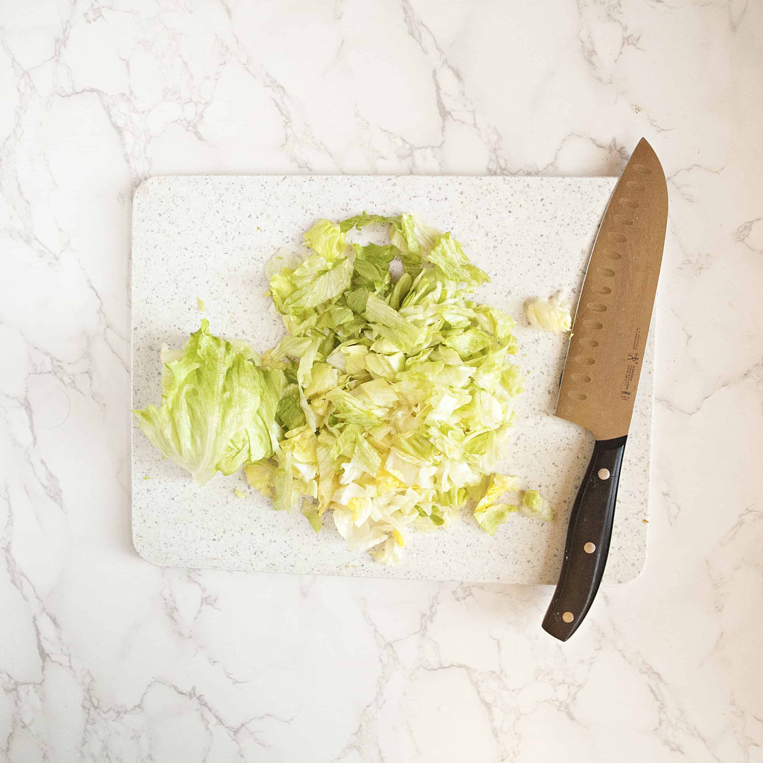 Chopped lettuce on a white cutting board with a chef's knife on a white marble background.