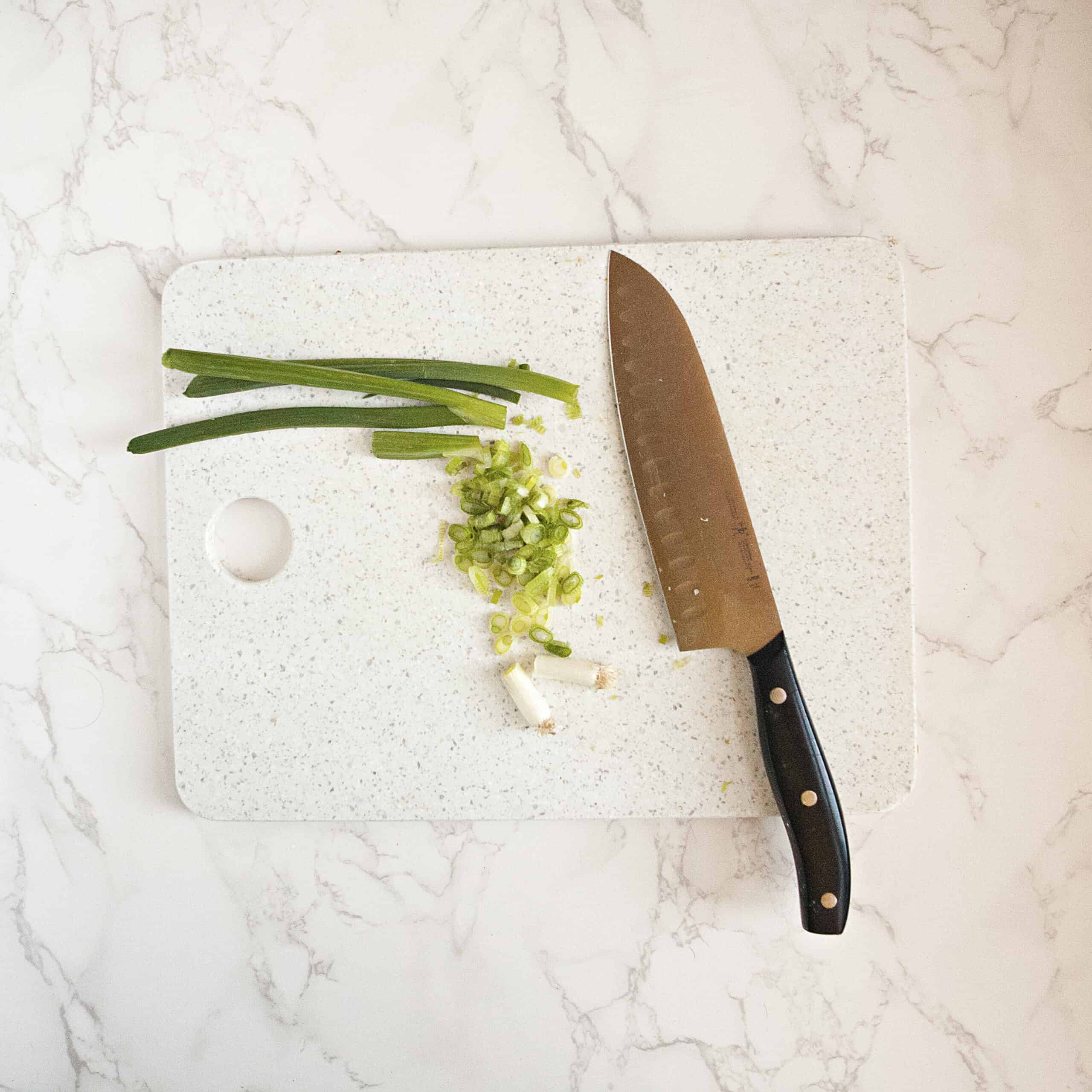 A chopped green onion on a white cutting board with a chef's knife on a white marble background.