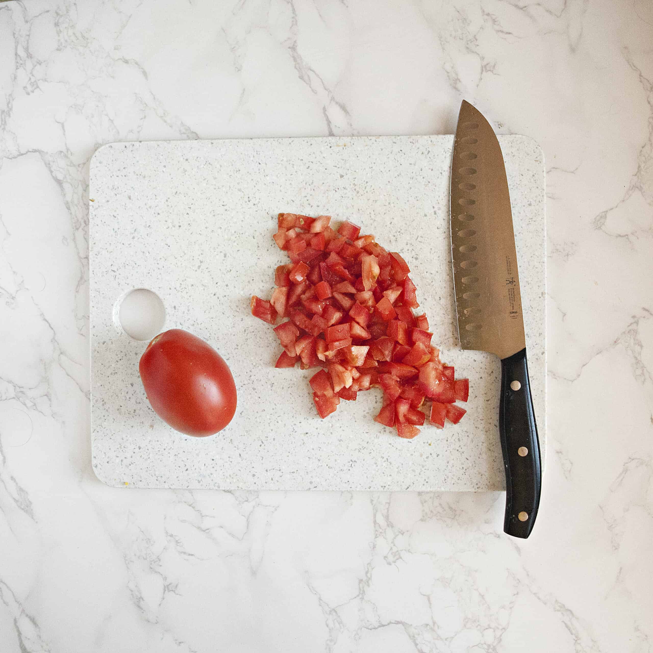 A Roma tomato and diced tomato pieces next to a chef's knife on a white cutting board on a white marble background.