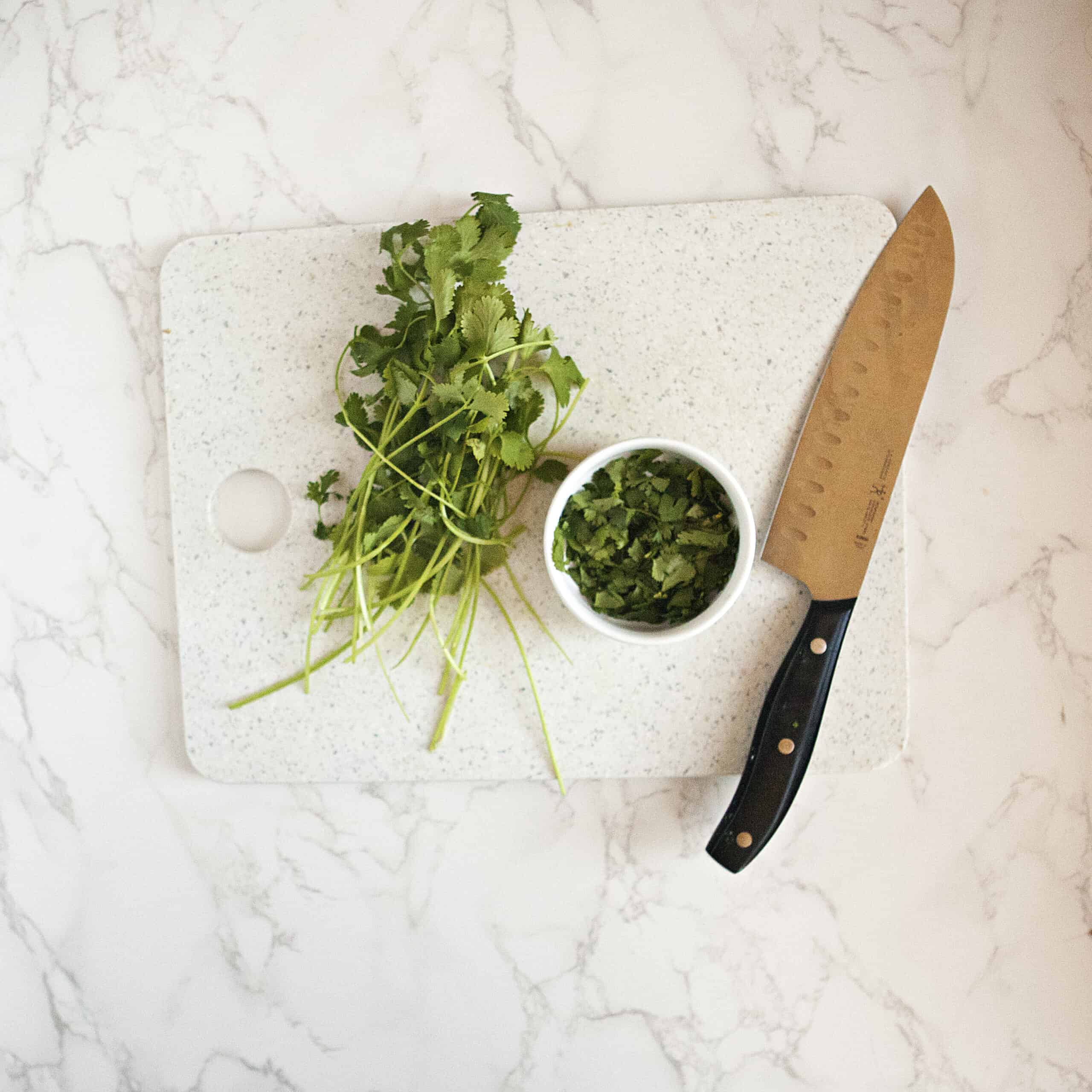 Fresh cilantro stems next to a small bowl containing chopped cilantro leaves and a chef's knife on a white cutting board, on a white marble background.