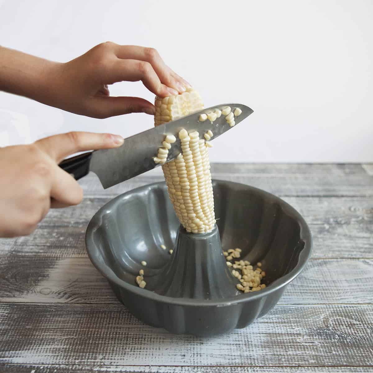 Two hands cutting corn off a cob with a knife.  The corn cob is inserted in the hole of a bundt pan.