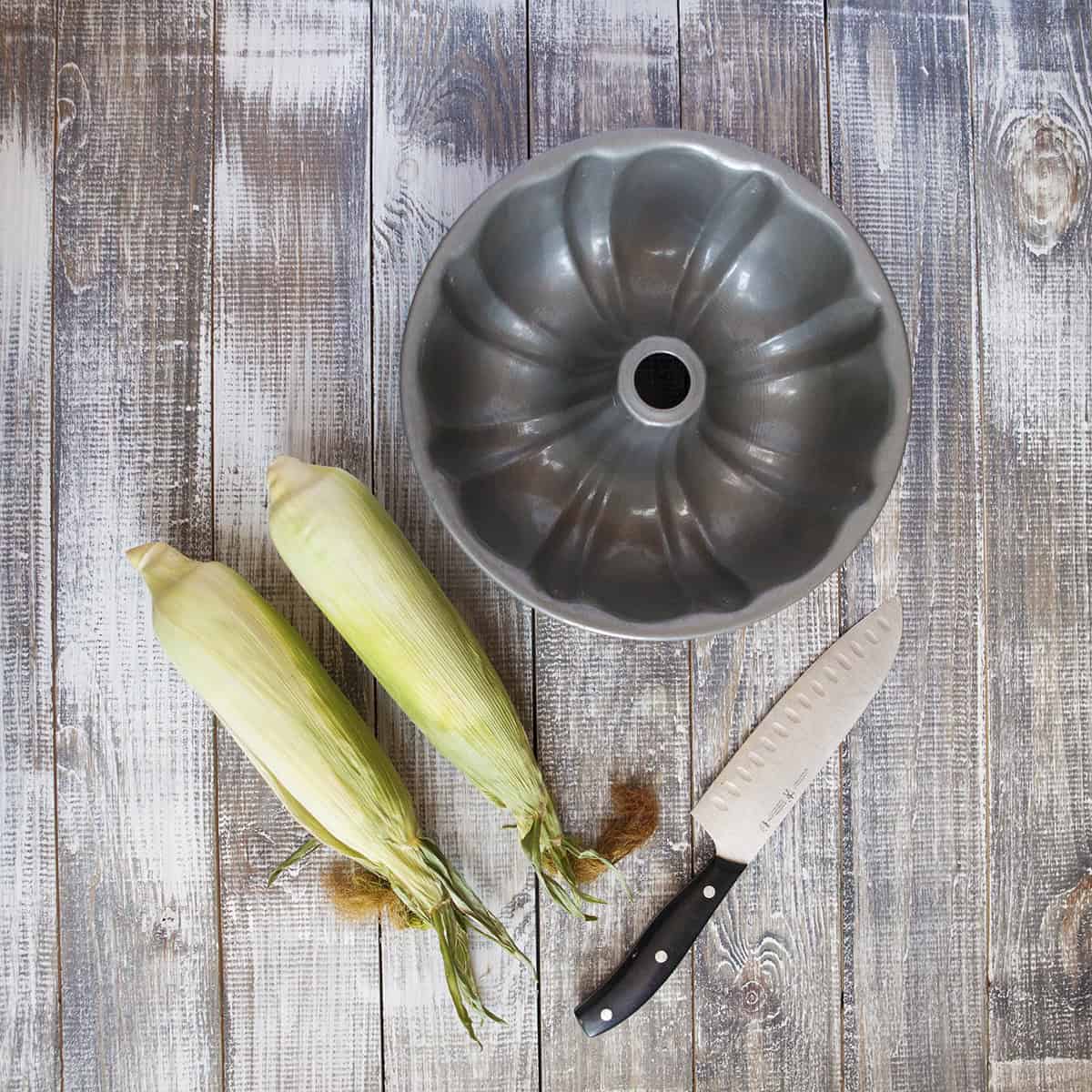 Two ears of corn next to a bundt cake pan and a chef's knife.