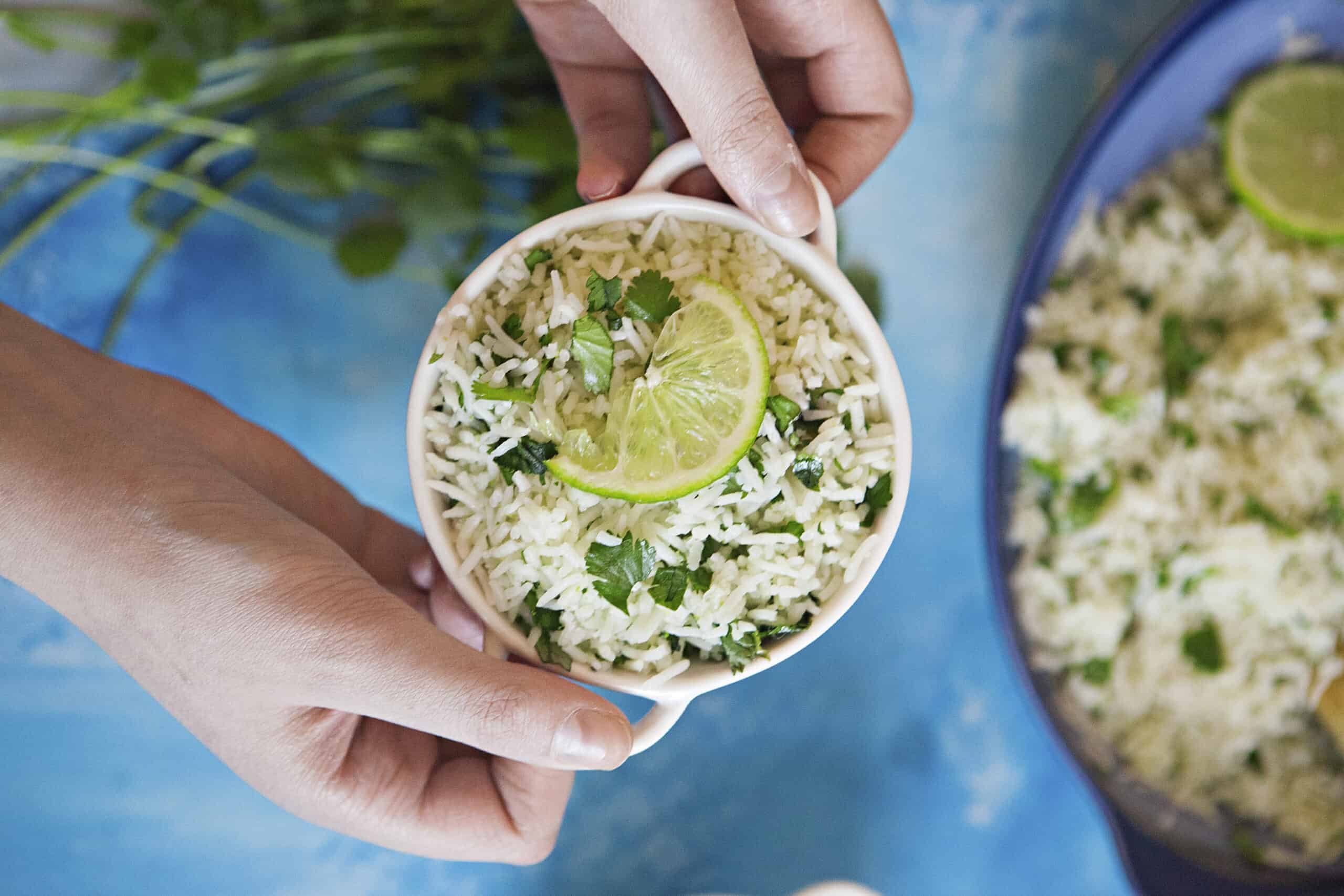 Fluffy cilantro-lime rice in a serving bowl being held by two hands over a blue background with fresh cilantro and a serving dish containing more rice.