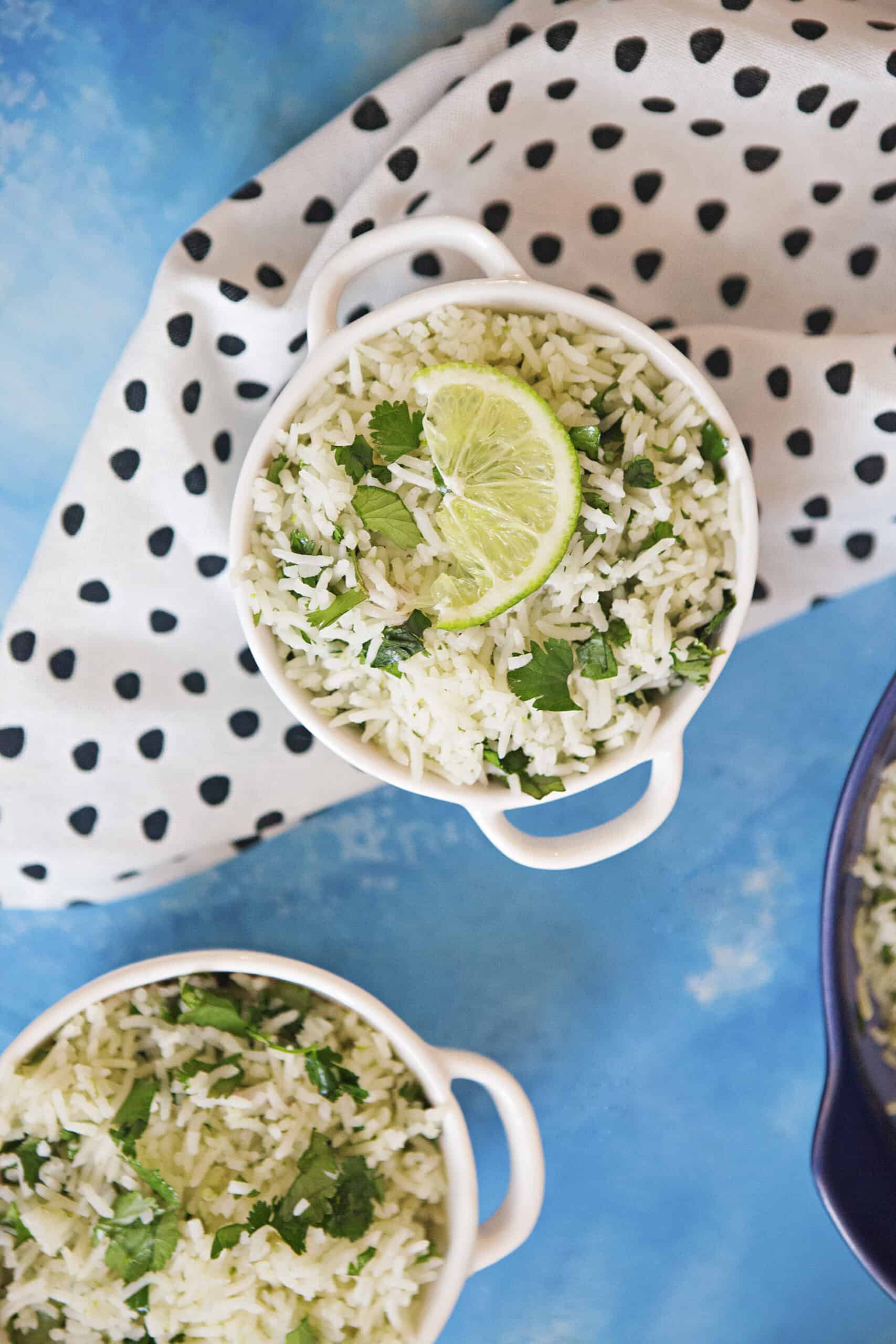 Two small bowls of cilantro-lime rice with a black and white dotted napkin alongside a blue serving dish containing more cilantro-lime rice and a wooden spoon on a blue background.