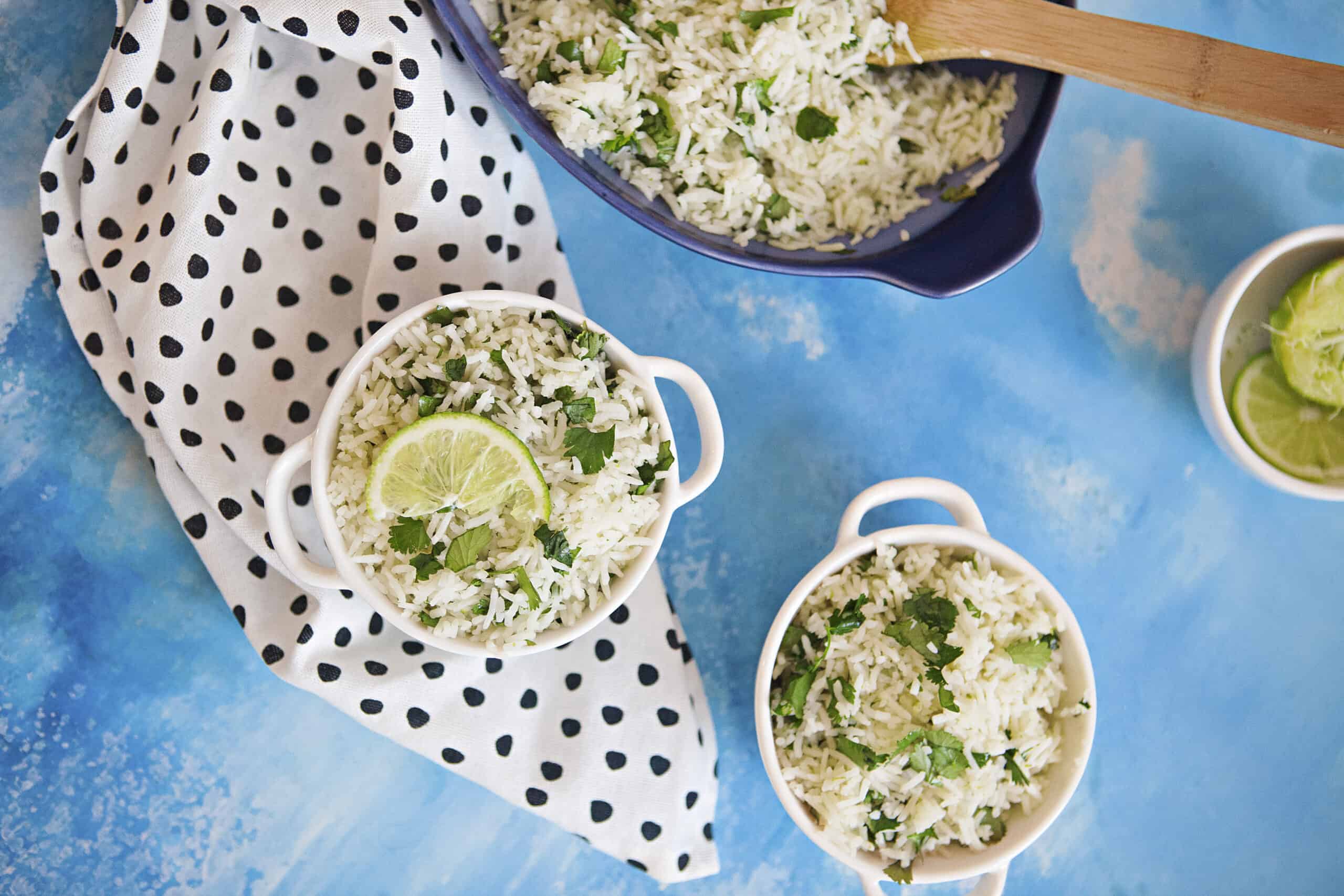 Two small bowls of cilantro-lime rice with a black and white dotted napkin alongside a blue serving dish containing more cilantro-lime rice and a wooden spoon on a blue background.