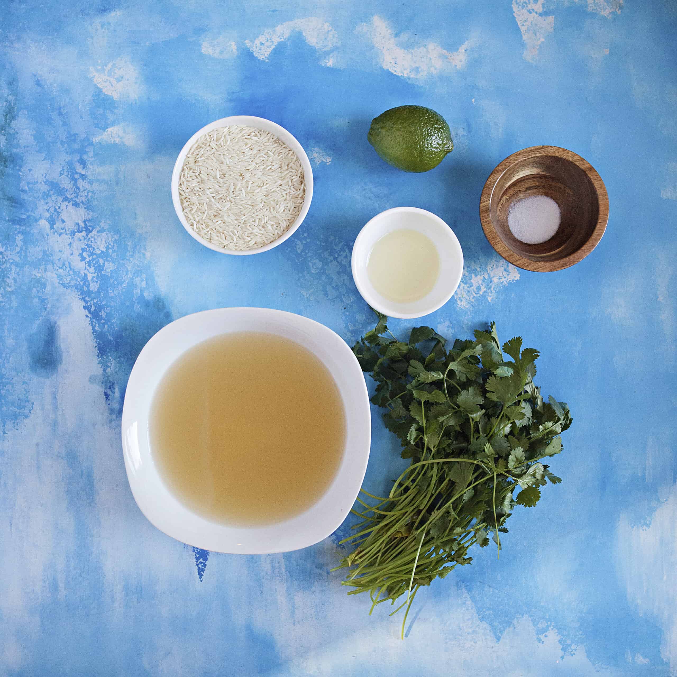 Ingredients for cilantro-lime rice:  Chicken broth, long-grain rice, fresh cilantro, a lime, olive oil, and salt, on a blue background.