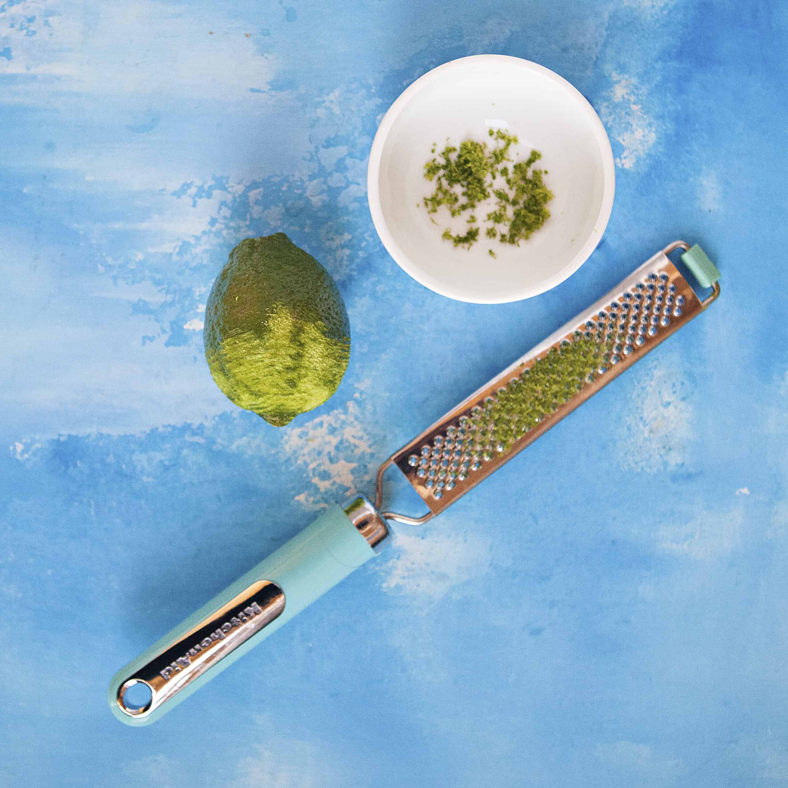 Partially zested lime alongside a microplane and a bowl containing the lime zest on a blue background.