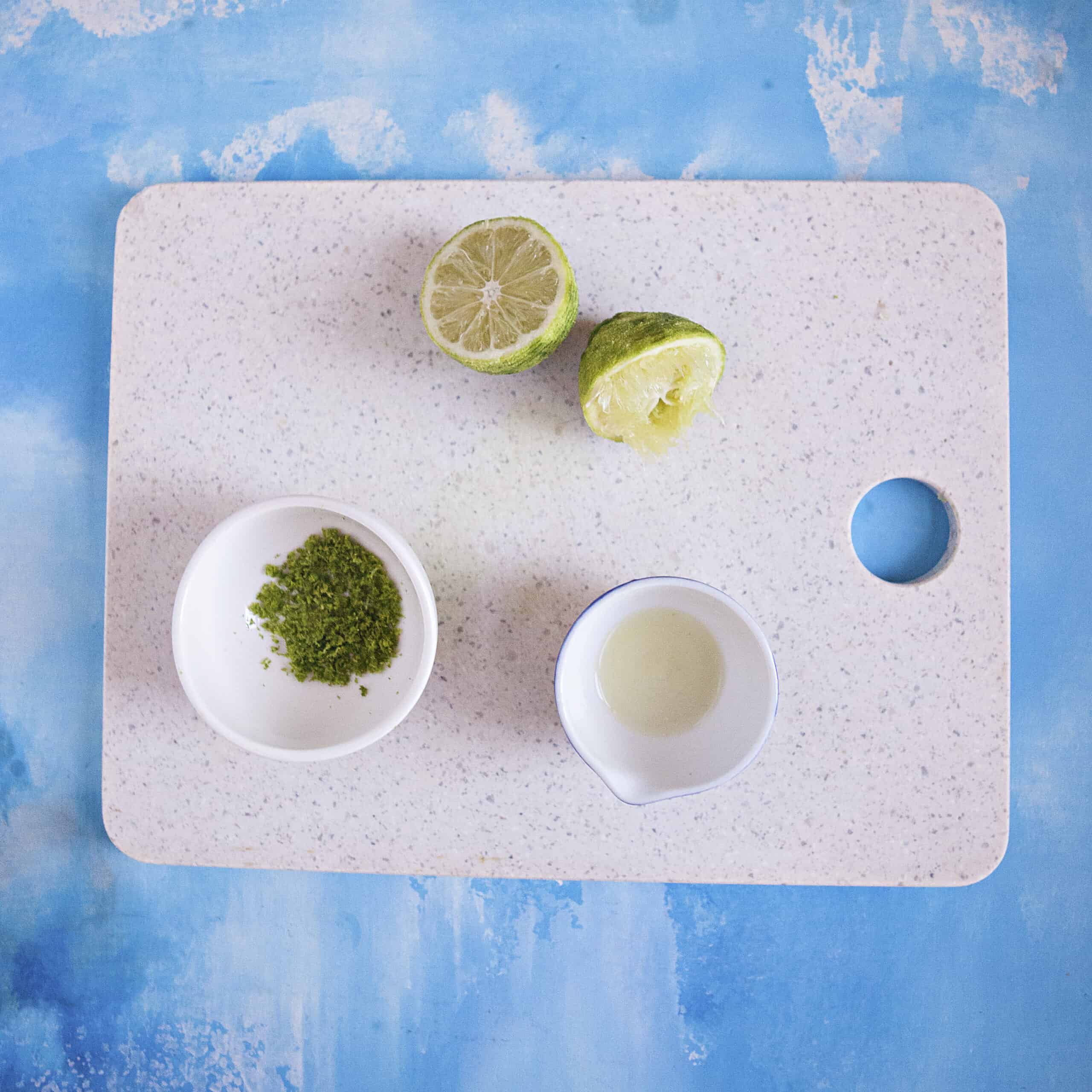 A sliced and juiced lime on a cutting board with small bowls containing lime zest and lime juice on a blue background.