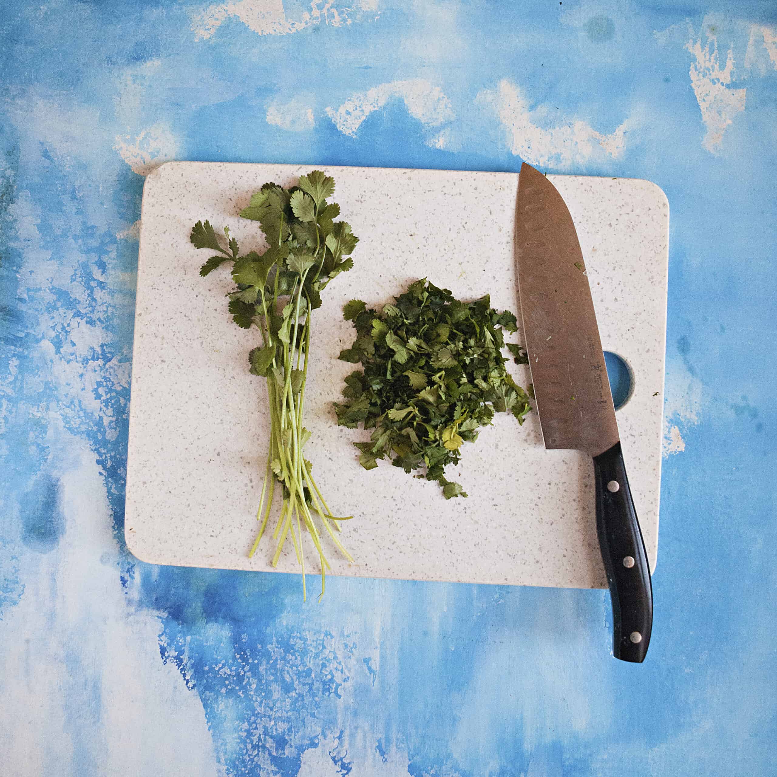 Fresh cilantro and chopped cilantro on a cutting board next to a chef's knife on a blue background.