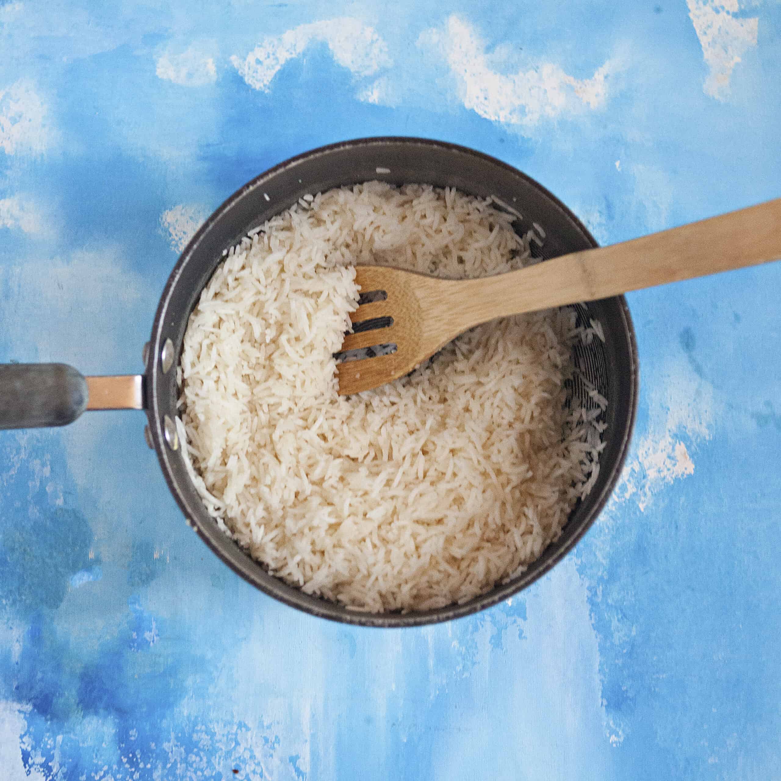 Cooked white rice in a pot with a wooden spoon on a blue background.