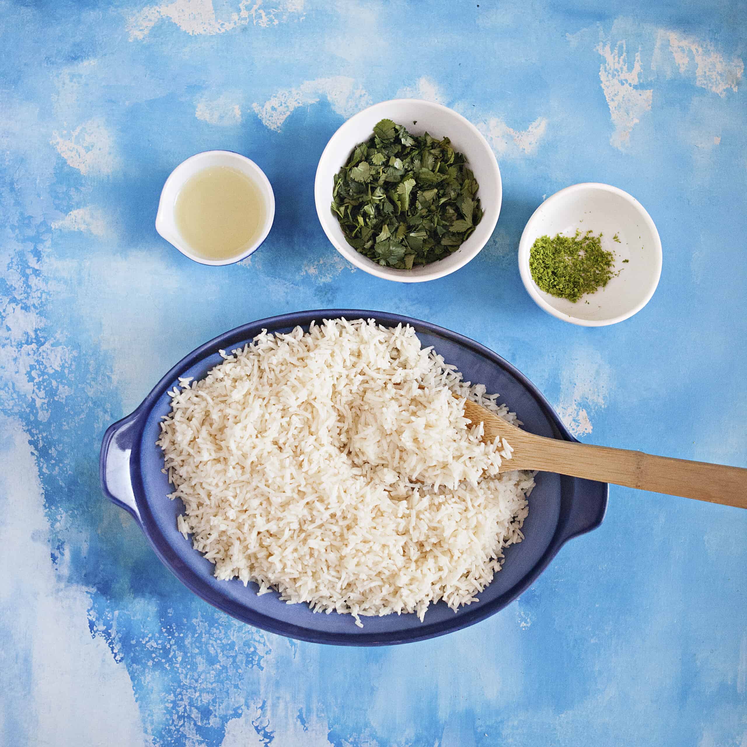 A blue serving dish of white rice with a wooden spoon next to 3 small bowls containing lime juice, chopped cilantro, and lime zest.