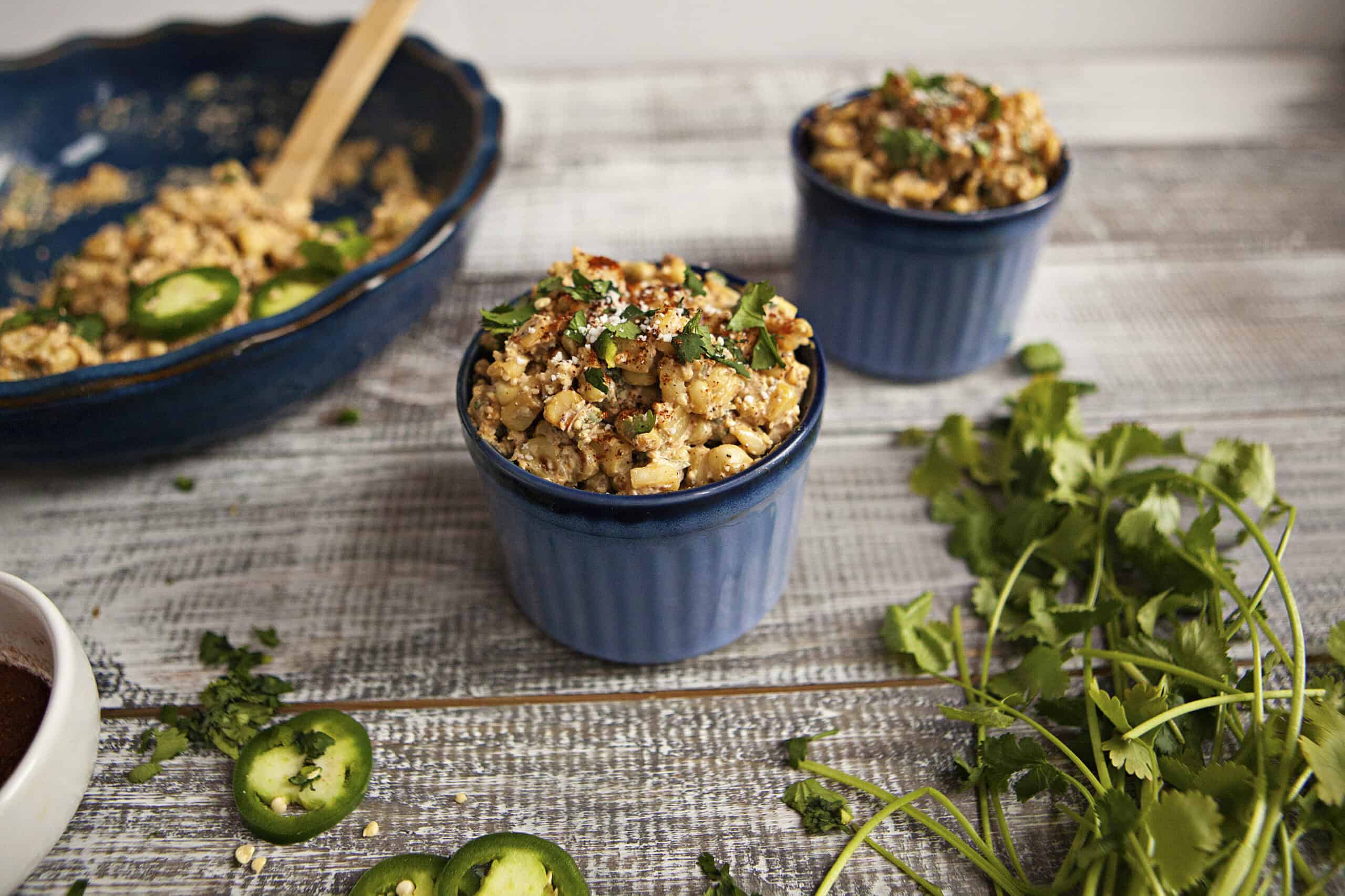 Two blue ramekins containing Mexican street corn next to a pie plate with more Mexican street corn, fresh cilantro, and slices of jalapeno pepper.