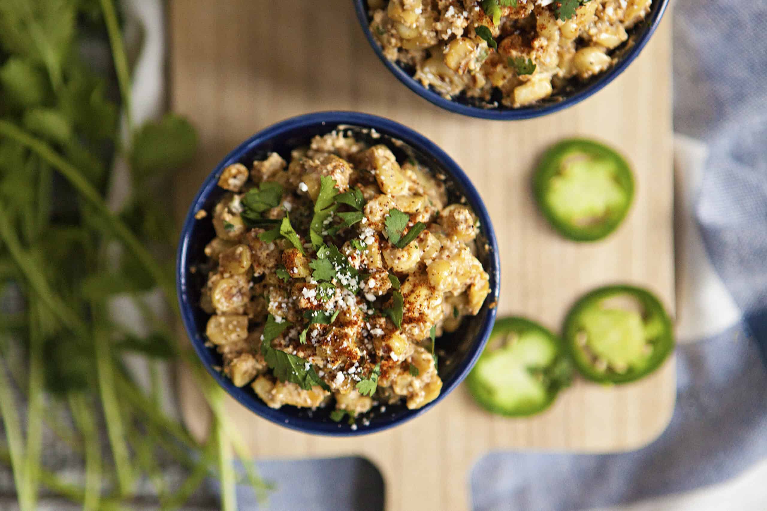 A blue ramekin holding Mexican street corn topped with chopped cilantro, chili powder, and Cotija cheese on a cutting board alongside more cilantro and slices of jalapeno pepper.