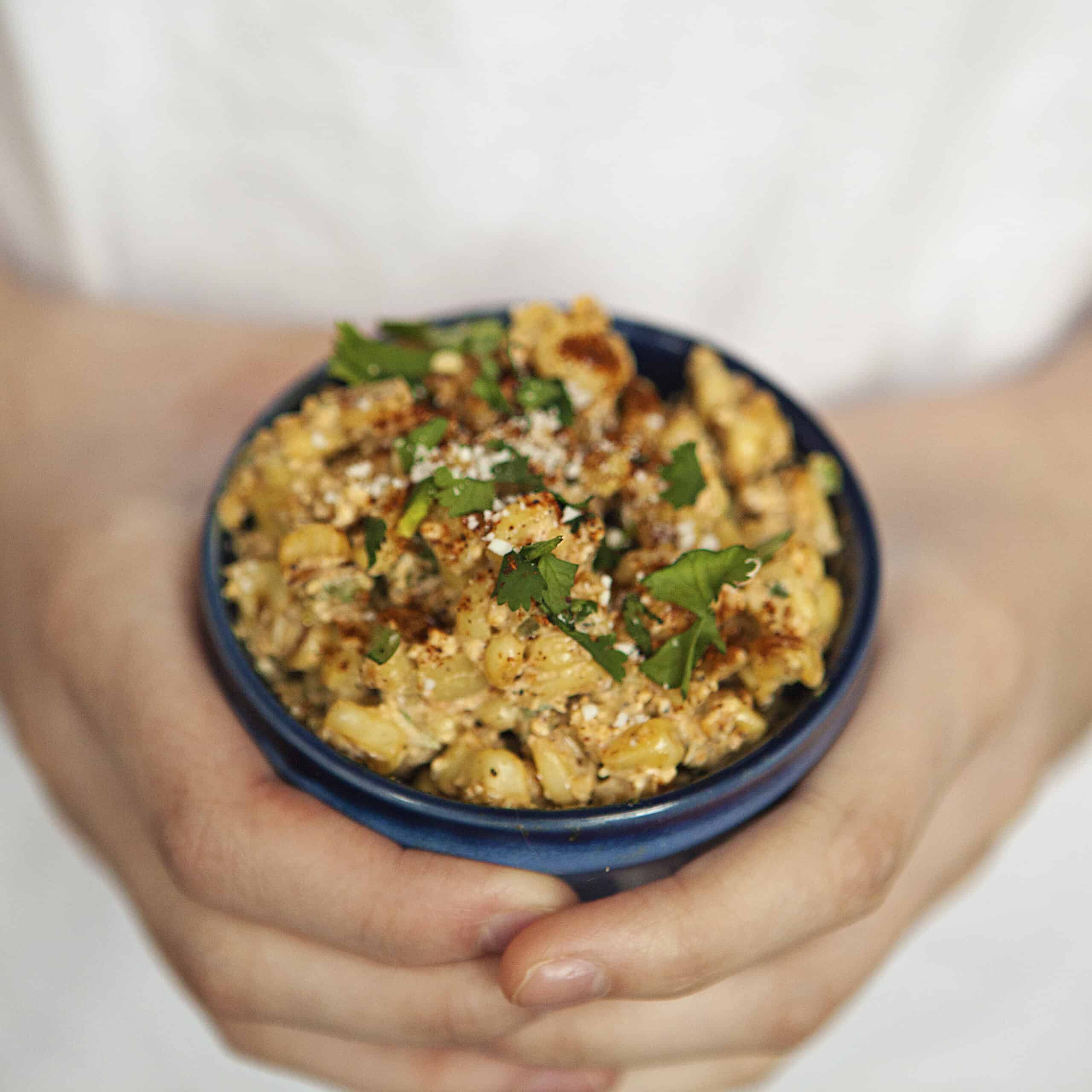 A cup of Mexican street corn topped with chopped cilantro, chili powder, and Cotija cheese being held by two hands.