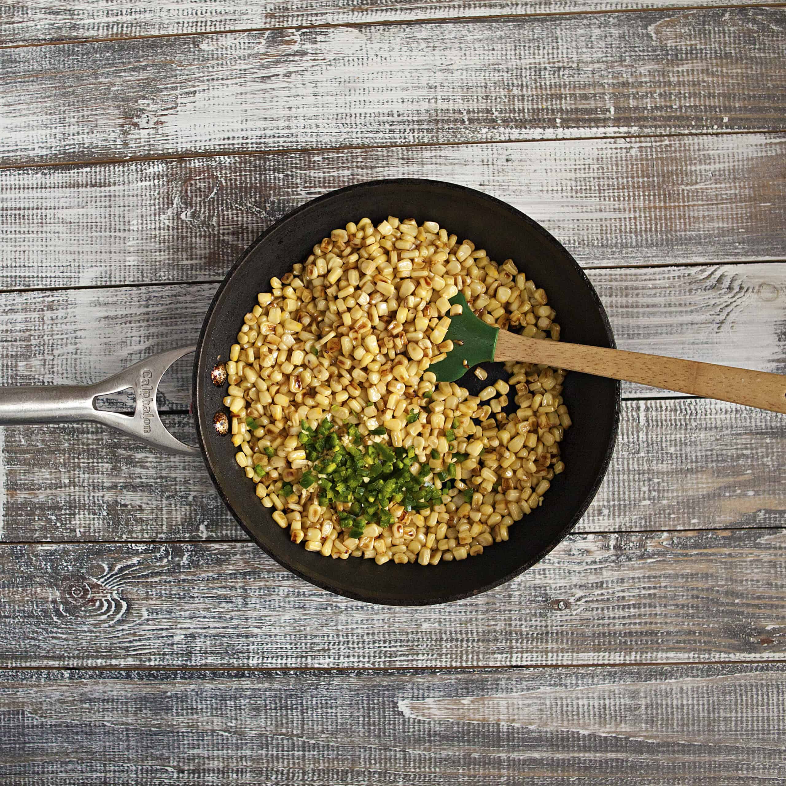 Corn kernels in a skillet with minced garlic and diced jalapeno with a wooden spoon.