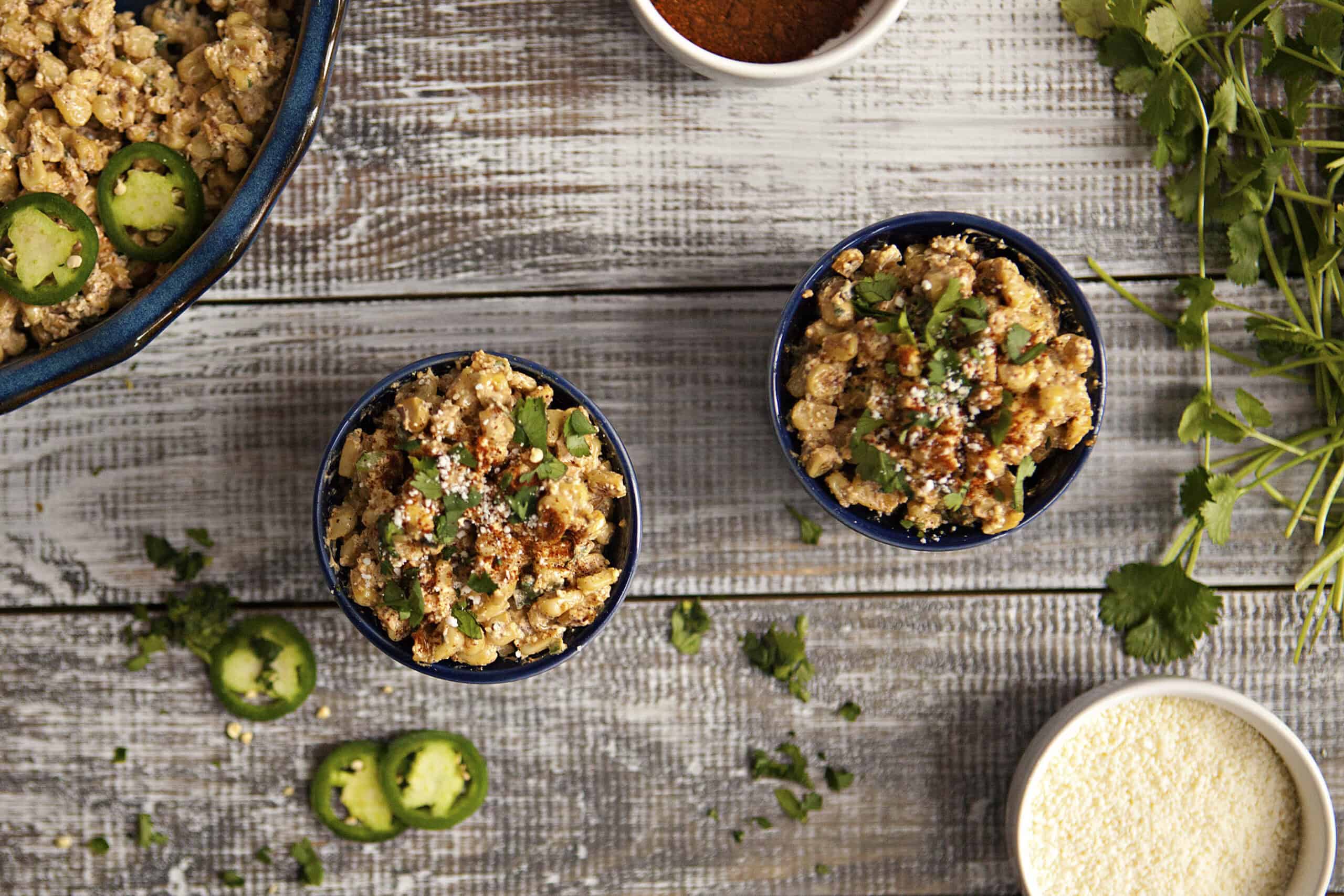 Mexican street corn (elote en vaso or esquites) in blue ramekins topped with chopped cilantro, chili powder, and Cotija cheese, next to more chopped cilantro and slices of jalapeno.