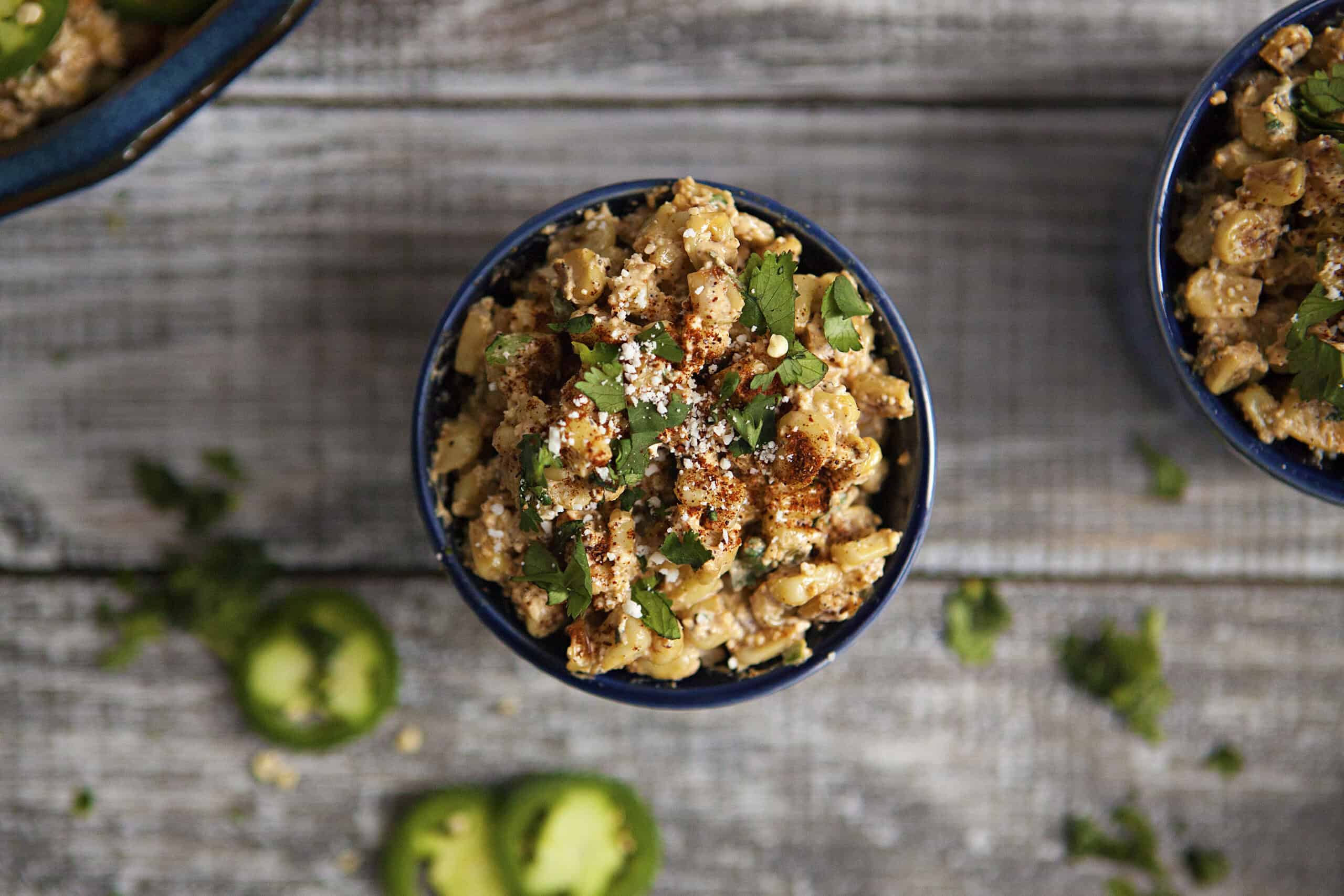 Mexican street corn (elote en vaso or esquites) in a blue ramekin topped with chopped cilantro, chili powder, and Cotija cheese, next to more chopped cilantro and slices of jalapeno.