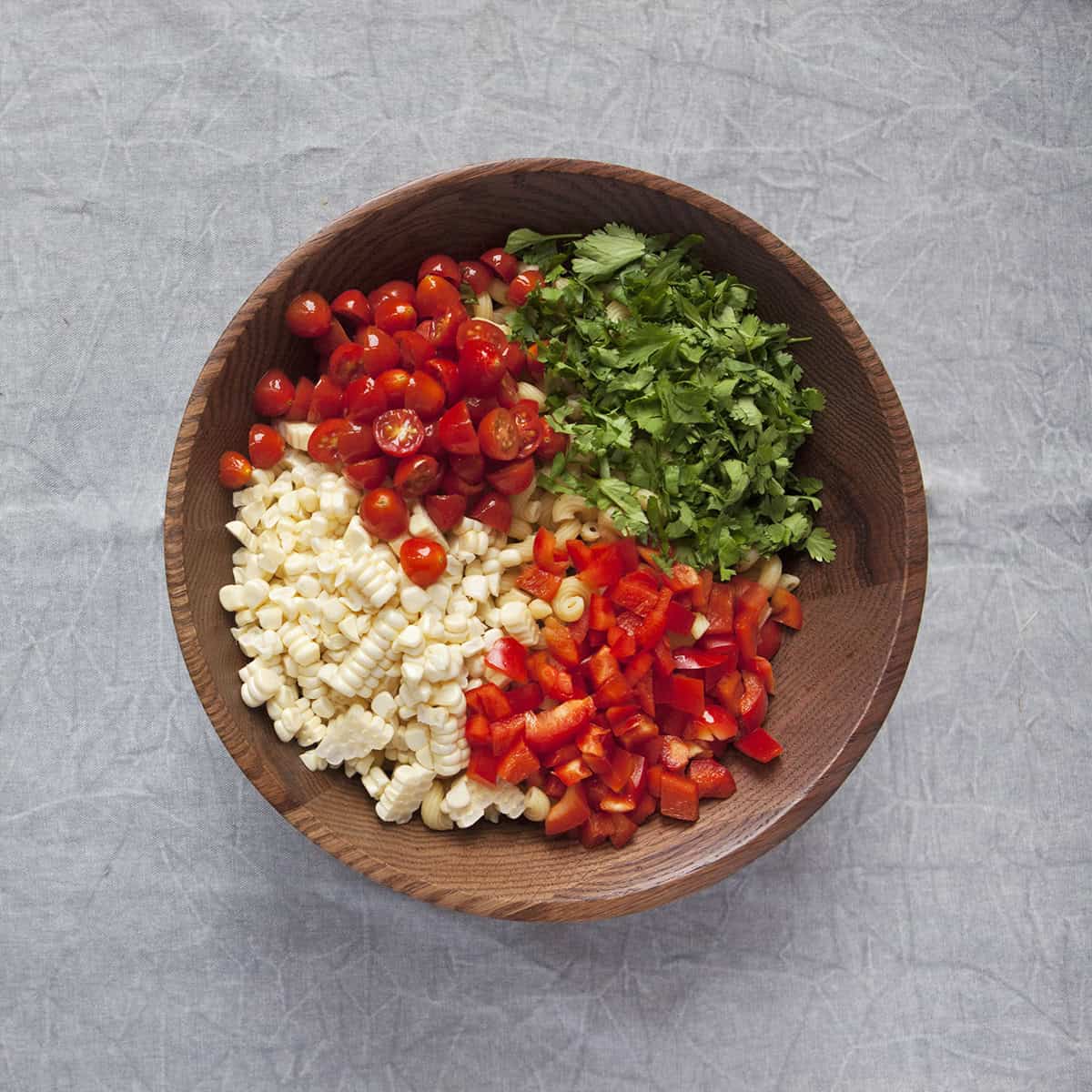 Chopped cherry tomatoes, red bell pepper, corn kernels, cilantro, and fusilli pasta in a wooden salad bowl on a gray background.