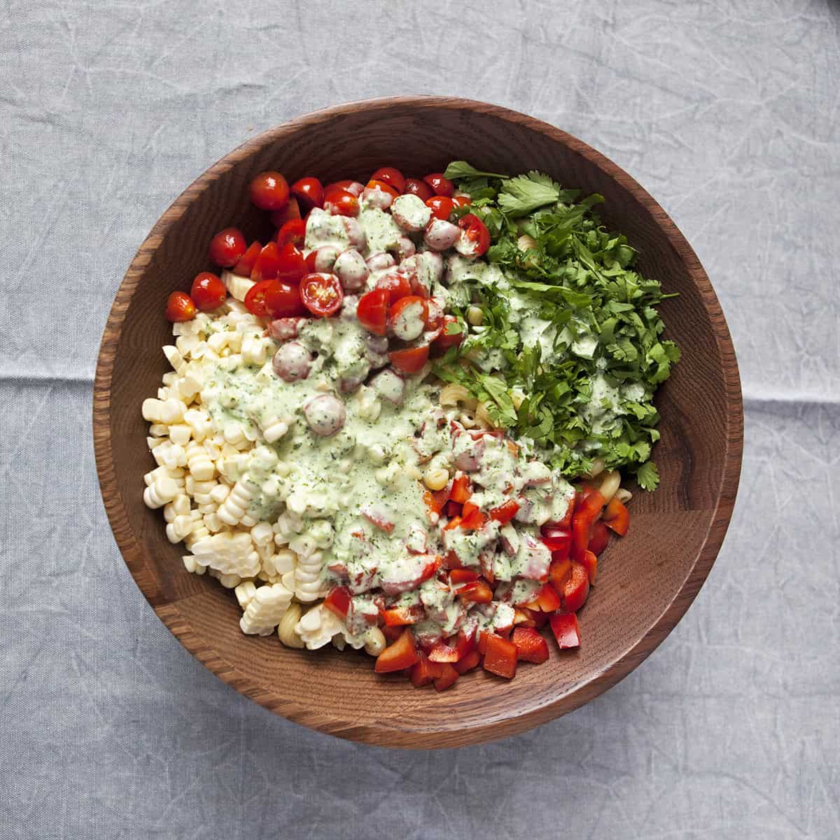 Chopped cherry tomatoes, cilantro, red bell pepper, corn kernels, and fusilli pasta topped with tomatillo ranch salad dressing in a wooden salad bowl on a gray background.