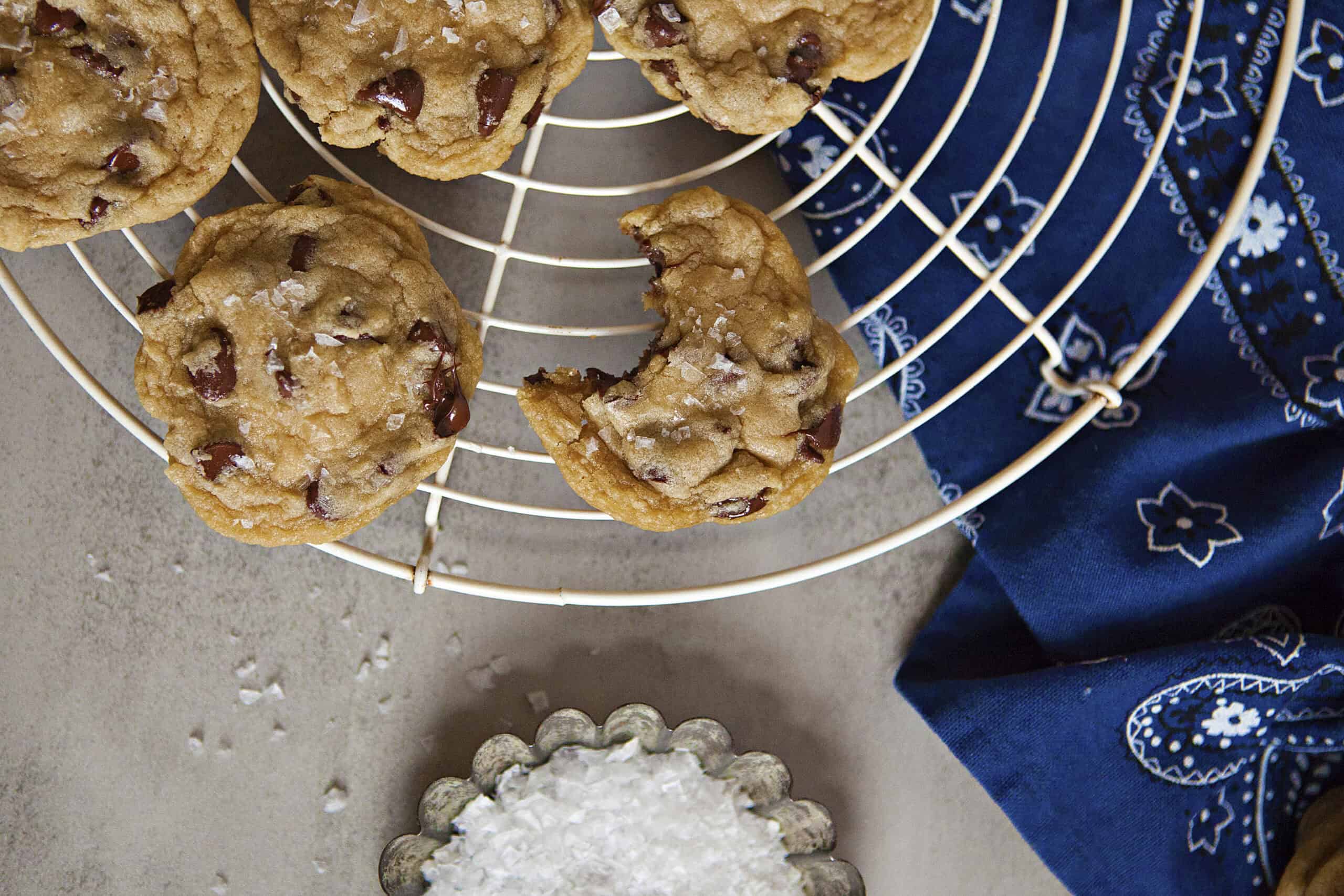 Soft chocolate chip cookies with sea salt on on white plates with a blue bandanna pattern kitchen towel on a gray background.