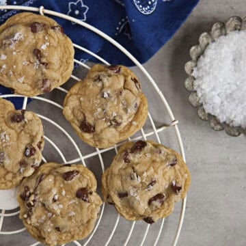 Soft chocolate chip cookies with sea salt on a cooling rack alongside a metal tart dish filled with sea salt flakes and a blue bandanna pattern kitchen towel on a gray background.