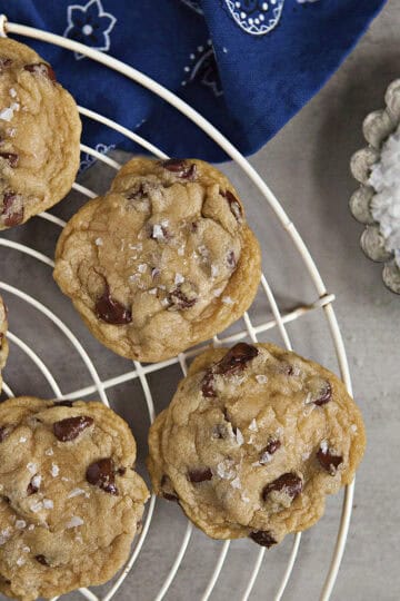 Soft chocolate chip cookies with sea salt on a cooling rack alongside a metal tart dish filled with sea salt flakes and a blue bandanna pattern kitchen towel on a gray background.