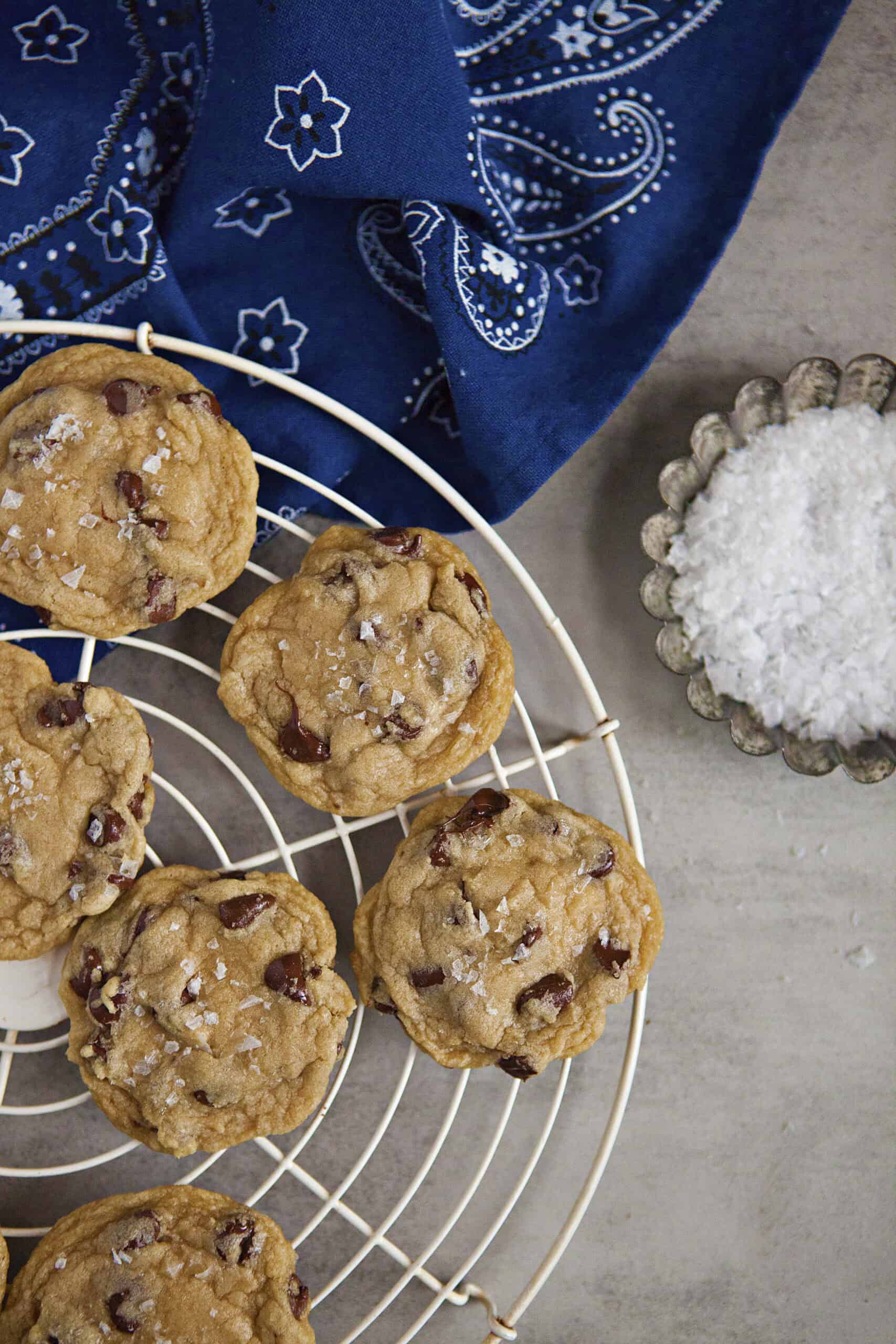 Soft chocolate chip cookies with sea salt on a cooling rack alongside a metal tart dish filled with sea salt flakes and a blue bandanna pattern kitchen towel on a gray background.