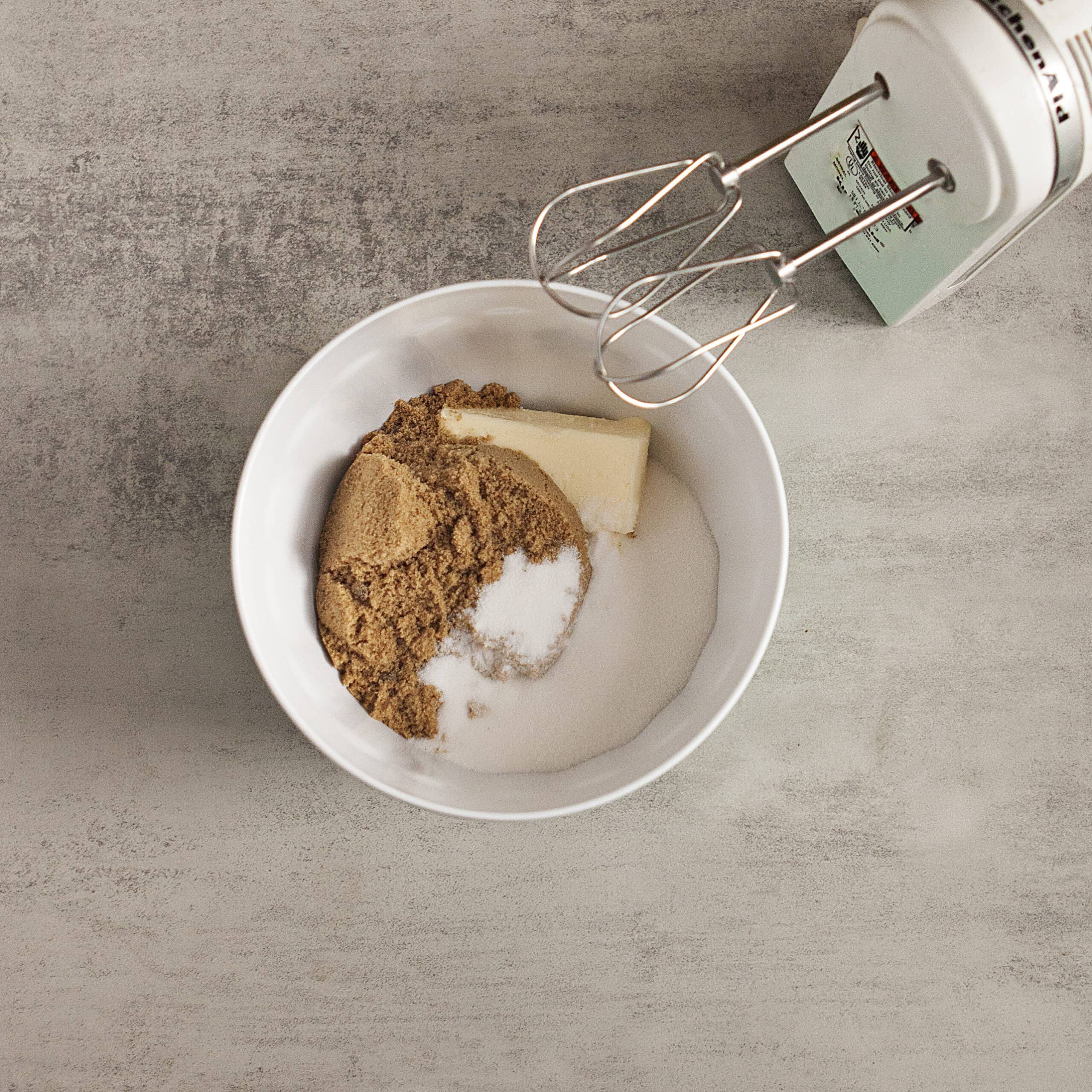 Butter, granulated sugar, and brown sugar in a mixing bowl alongside a handheld electric mixer on a gray background.