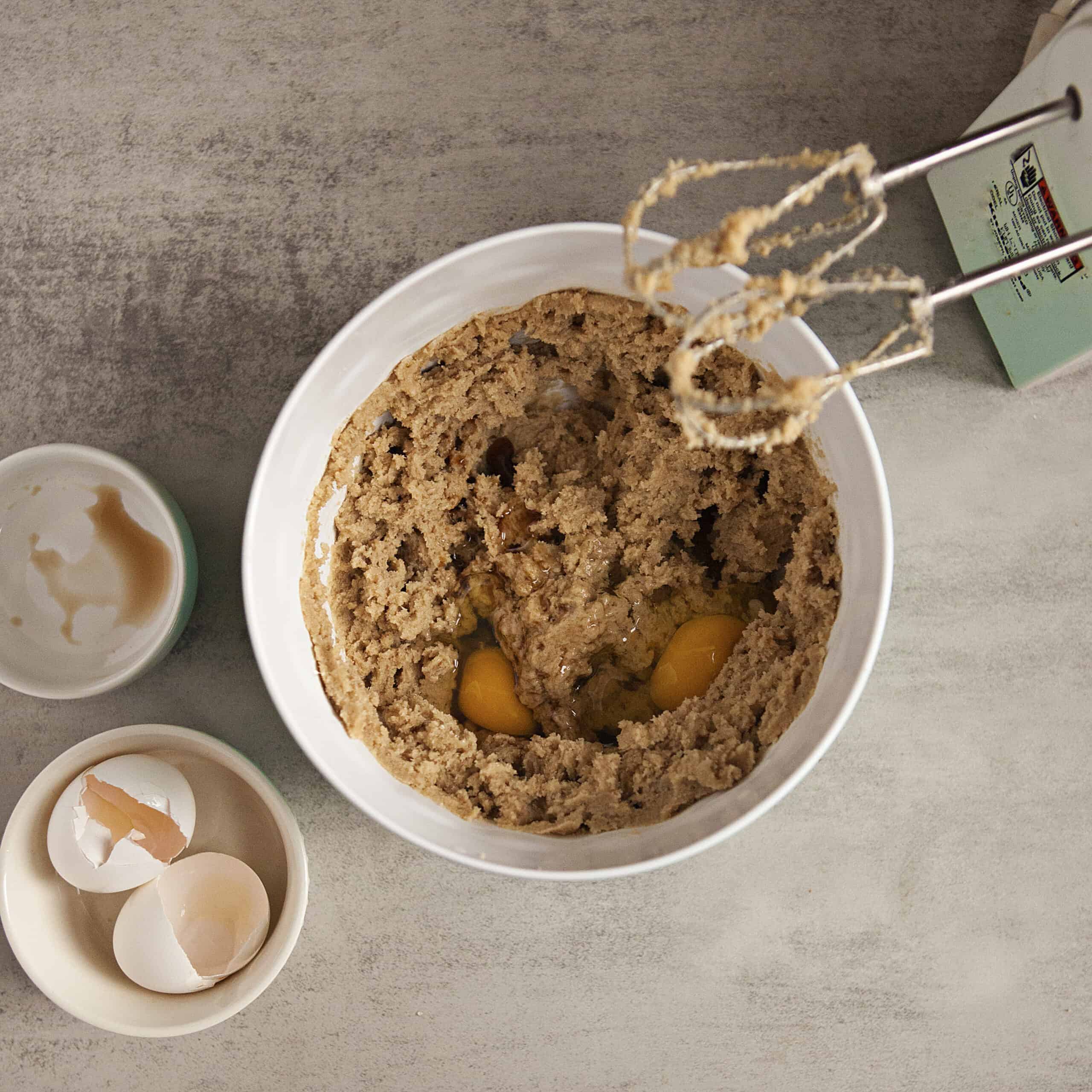 Cookie dough with eggs and vanilla being mixed in with an electric mixer next to small bowls containing vanilla residue and eggshells on a gray background.