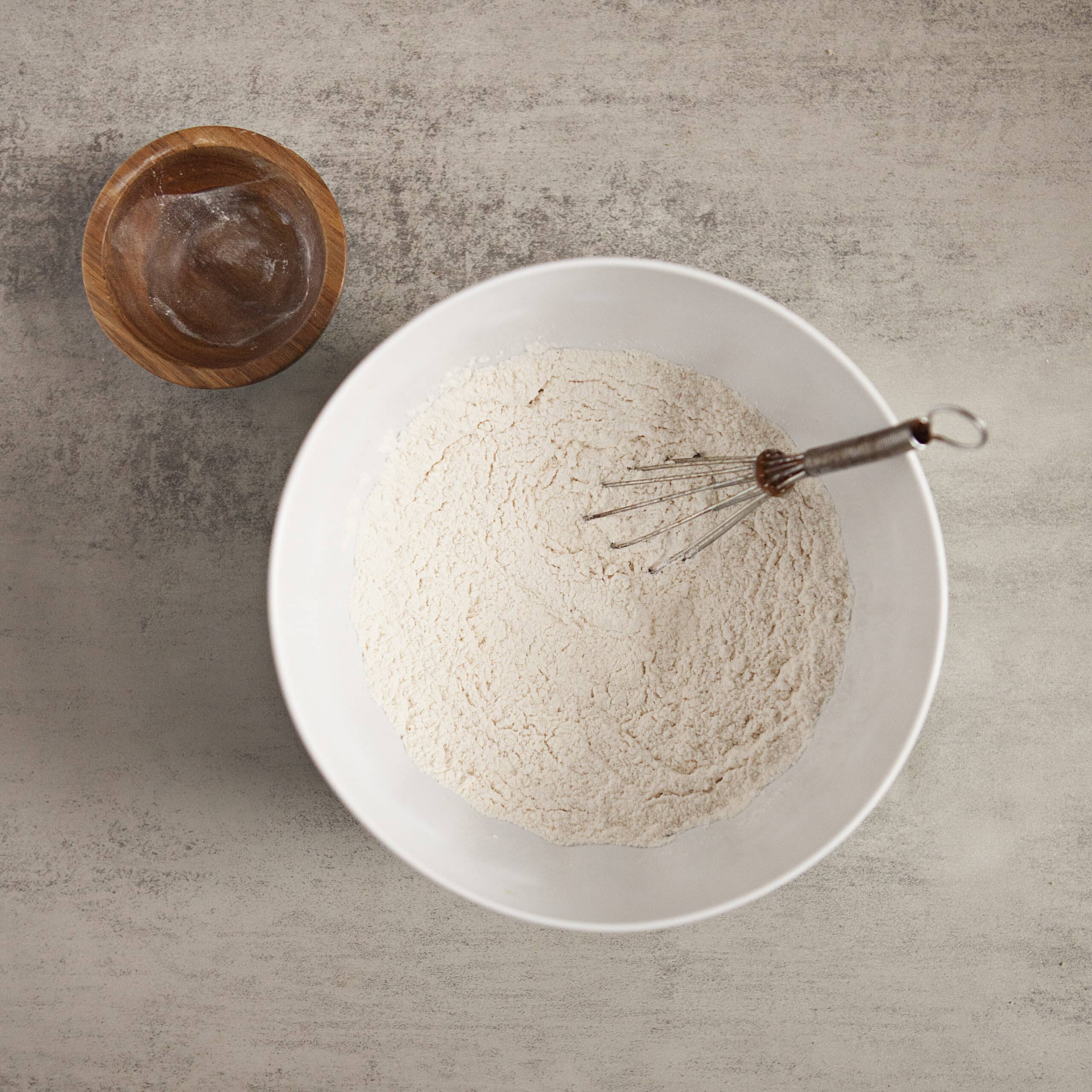 Dry ingredients being whisked together in a mixing bowl alongside a small wooden bowl with baking powder residue on a gray background.