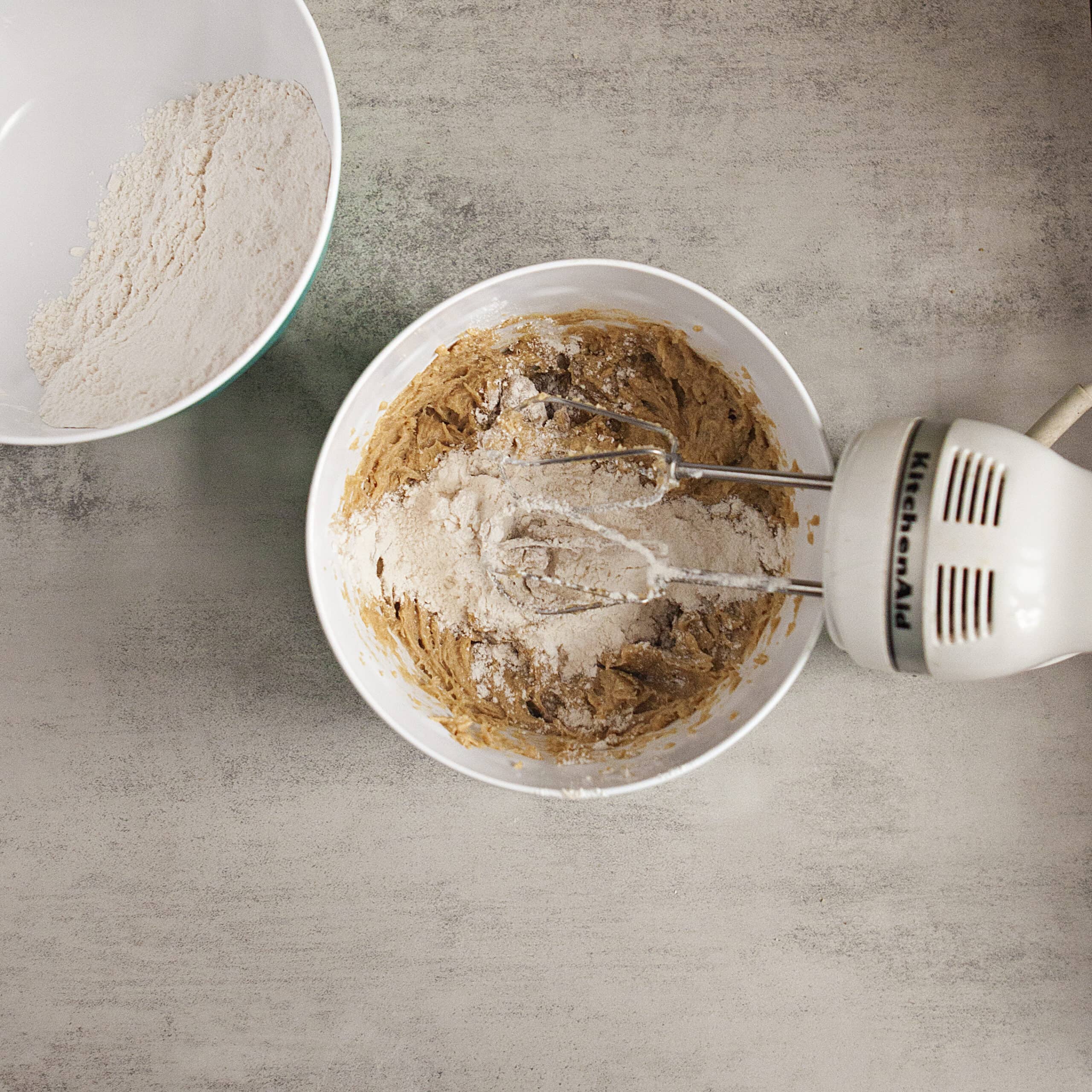 Cookie dough in a mixing bowl with flour being mixed in with an electric mixer next to another bowl containing more flour on a gray background.