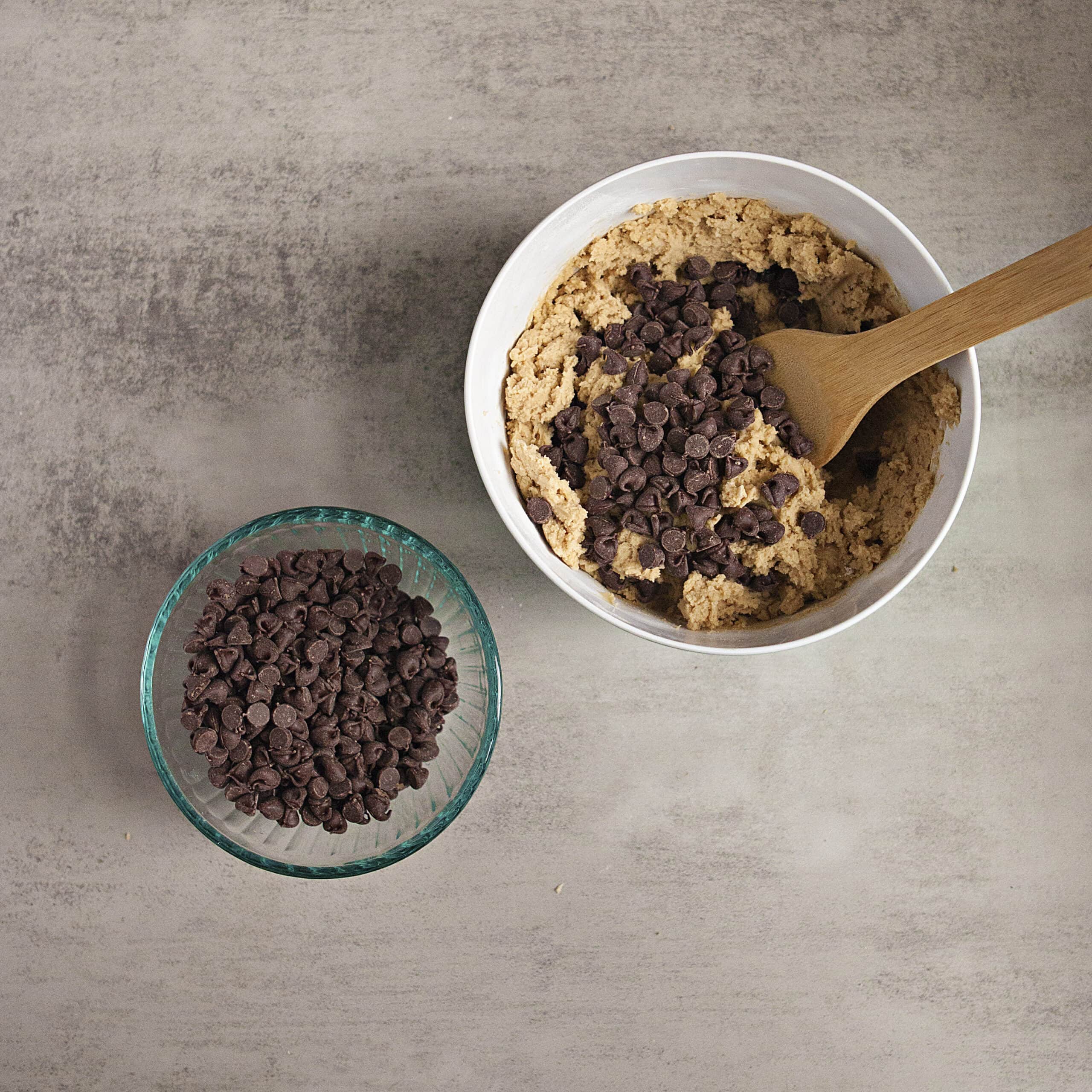 Cookie dough with chocolate chips being mixed in a mixing bowl with a wooden spoon next to a glass bowl containing more chocolate chips on a gray background.