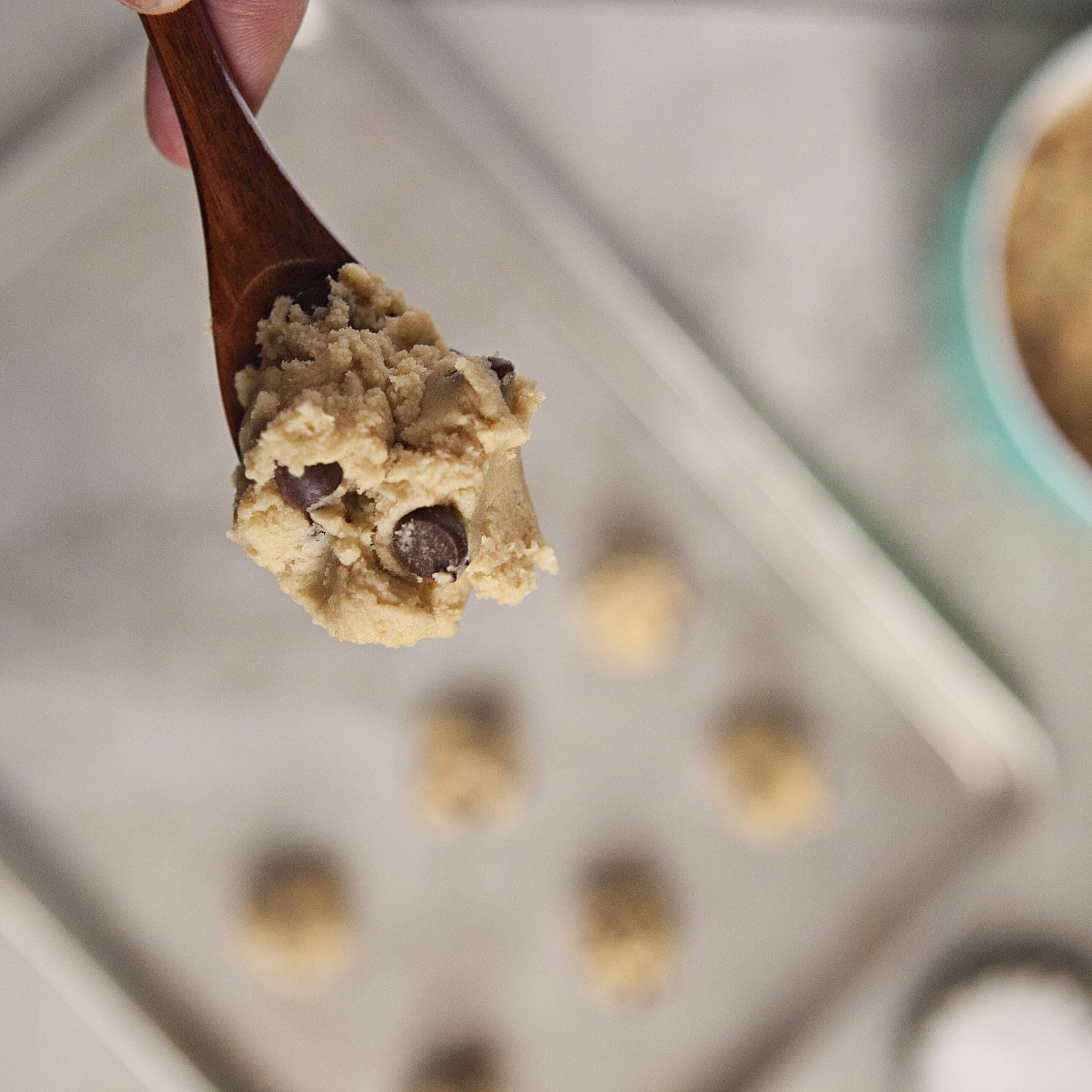 A spoonful of cookie dough in a small wooden spoon above a cookie sheet with more cookie dough blobs on a gray background.