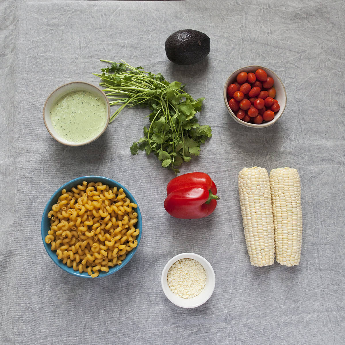 Ingredients for zesty tomatillo ranch pasta salad:  Fusilli pasta, corn, cherry tomatoes, avocado, cilantro, jalapeno ranch salad dressing, Cotija cheese, red bell pepper, on a gray background.