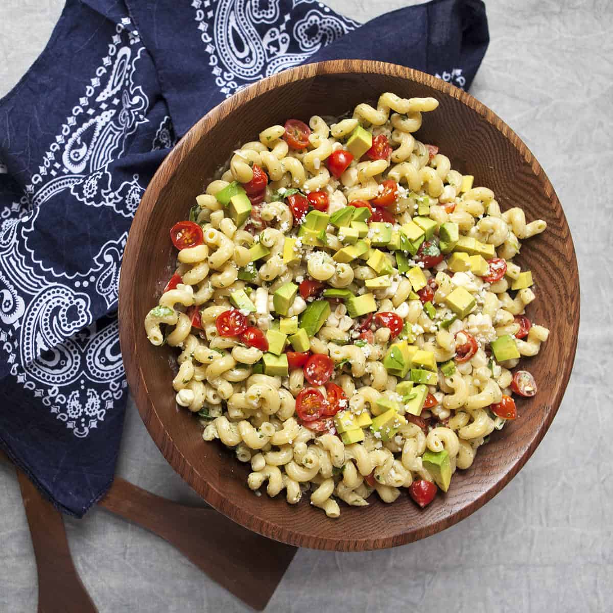 Zesty tomatillo ranch pasta salad with corn, bell peppers, avocado, cherry tomatoes, and cilantro in a large wooden salad bowl next to a navy blue handkerchief-style napkin and salad serving utensils on a gray background.