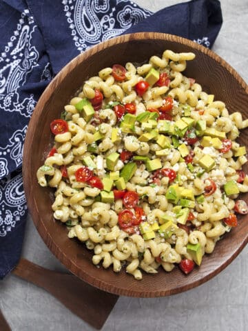 Zesty tomatillo ranch pasta salad with corn, bell peppers, avocado, cherry tomatoes, and cilantro in a large wooden salad bowl next to a navy blue handkerchief-style napkin and salad serving utensils on a gray background.