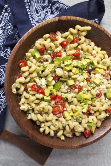 Zesty tomatillo ranch pasta salad with corn, bell peppers, avocado, cherry tomatoes, and cilantro in a large wooden salad bowl next to a navy blue handkerchief-style napkin and salad serving utensils on a gray background.