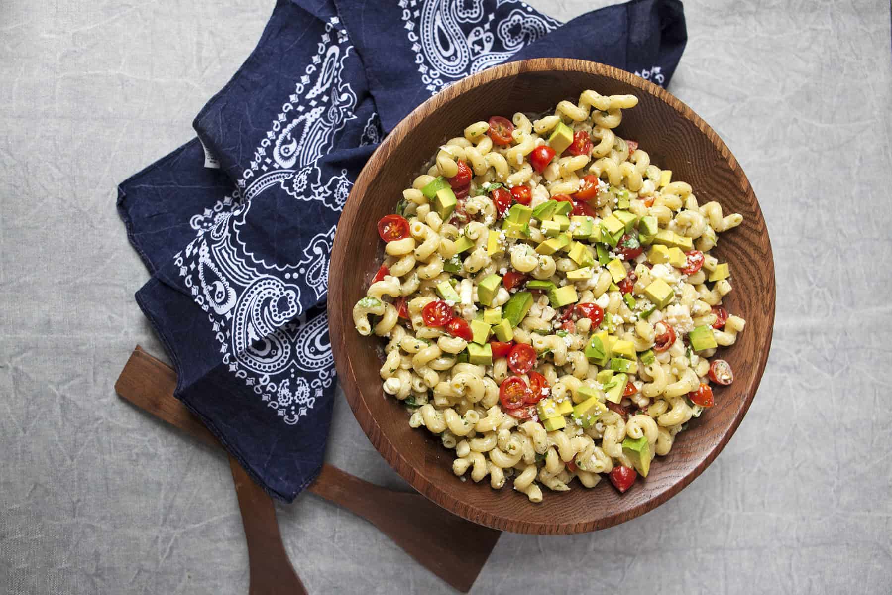 Zesty tomatillo ranch pasta salad with corn, bell peppers, avocado, cherry tomatoes, and cilantro in a large wooden salad bowl next to a navy blue handkerchief-style napkin and salad serving utensils on a gray background.