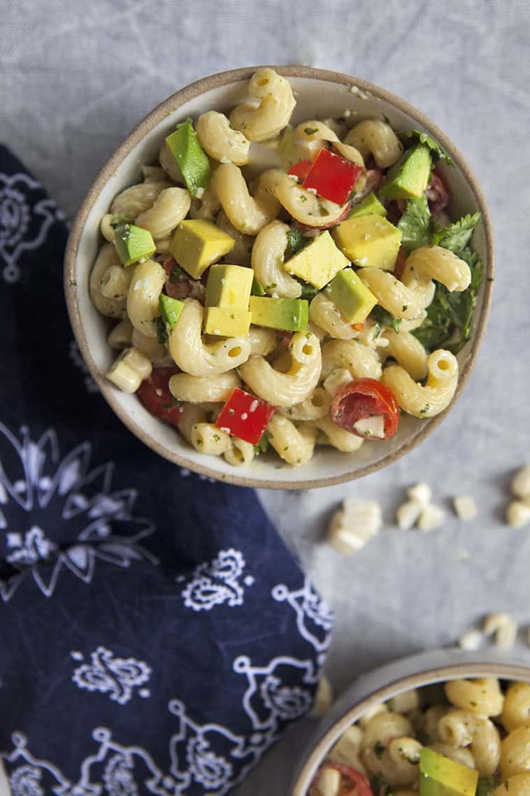 A white bowl of zesty tomatillo ranch pasta salad with corn, bell peppers, avocado, cherry tomatoes, and cilantro next to a navy blue handkerchief-style napkin on a gray background.