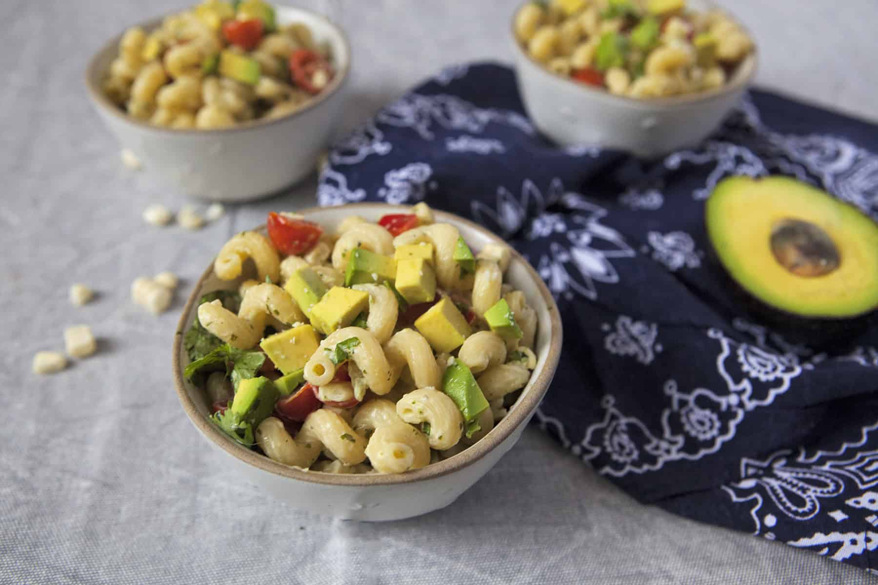 Zesty tomatillo ranch pasta salad with corn, bell peppers, avocado, cherry tomatoes, and cilantro in 3 small white bowls next to a navy blue handkerchief-style napkin on a gray background alongside half of an avocado and loose corn kernels.