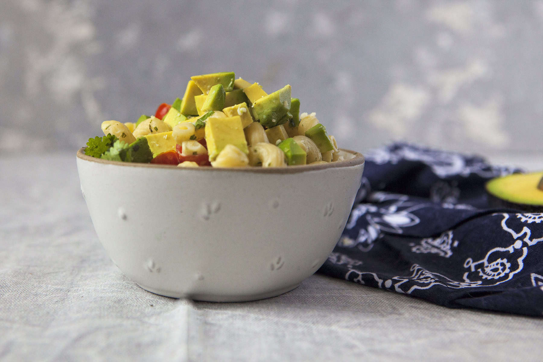 A white bowl of zesty tomatillo ranch pasta salad with corn, bell peppers, avocado, cherry tomatoes, and cilantro next to a navy blue handkerchief-style napkin on a gray background.