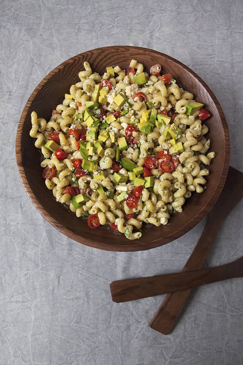 A large wooden salad bowl of zesty tomatillo ranch pasta salad with corn, bell peppers, avocado, cherry tomatoes, and cilantro next to wooden salad tongs on a gray background.