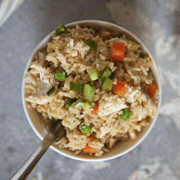 A bowl of chicken fried rice with egg, peas, carrots, and green onion on a gray background.