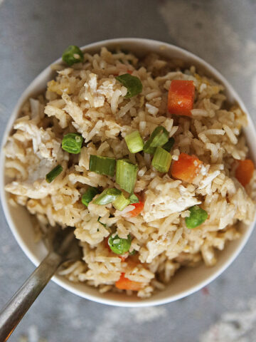 A bowl of chicken fried rice with egg, peas, carrots, and green onion on a gray background.