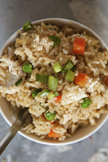 A bowl of chicken fried rice with egg, peas, carrots, and green onion on a gray background.