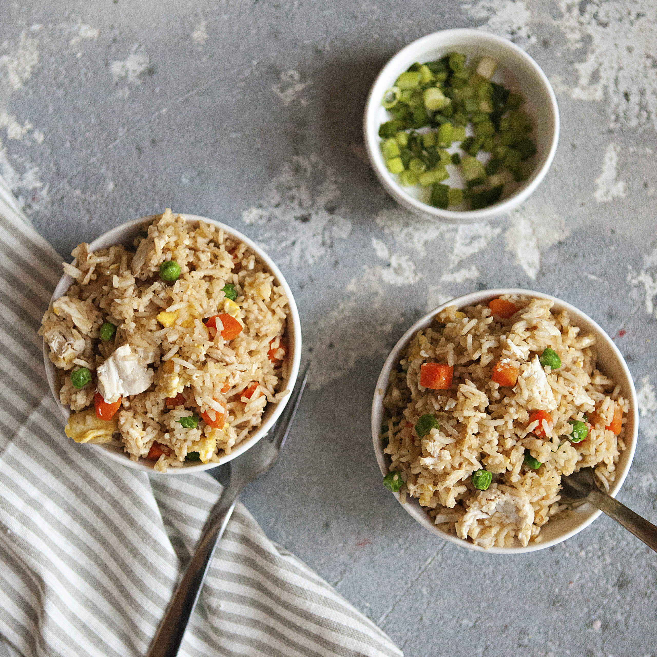 Two bowls of chicken fried rice with eggs, peas, carrots, and green onions next to a tiny bowl of chopped green onions and a white and gray striped napkin on a gray background.