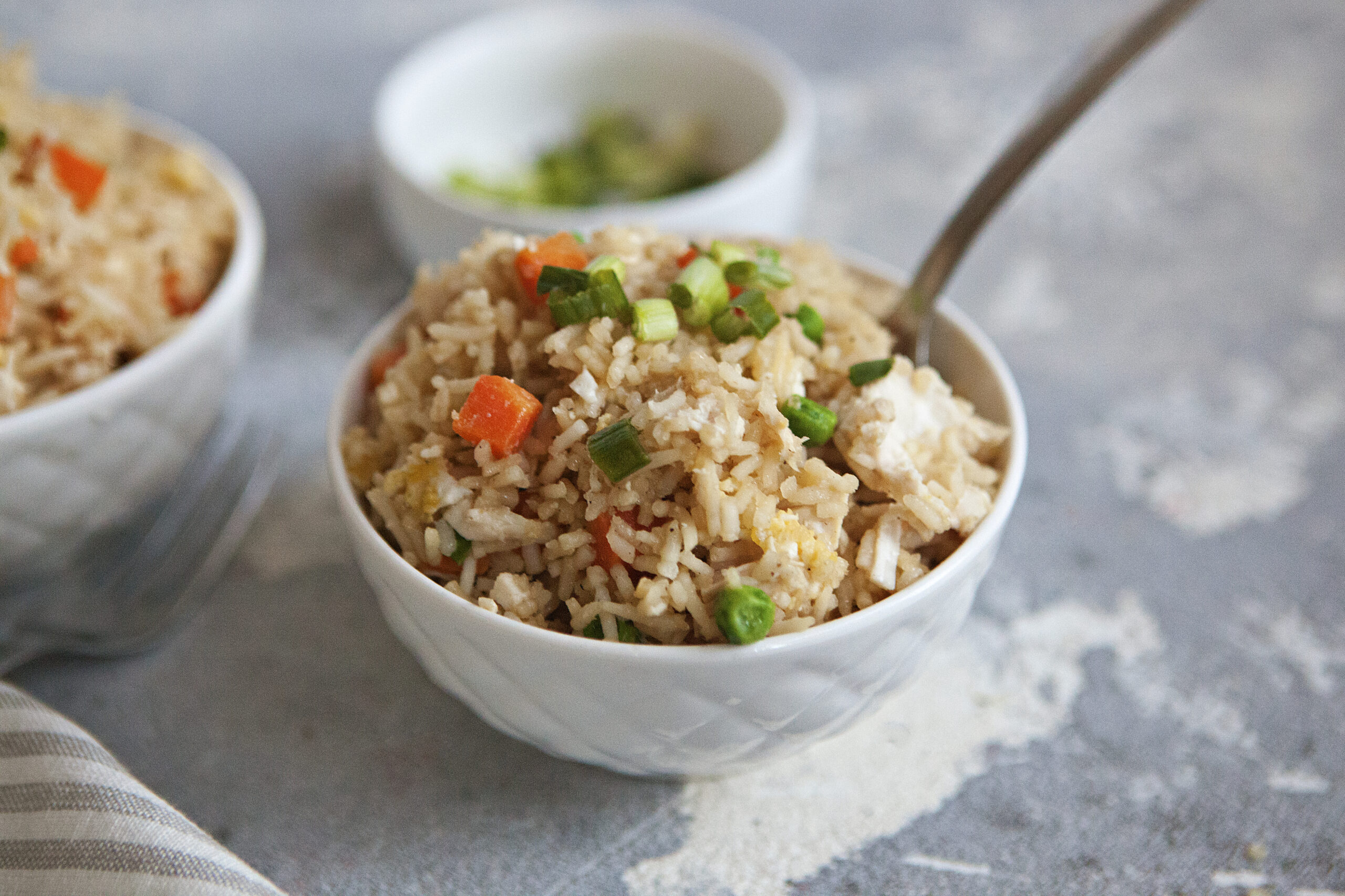 A bowl of chicken fried rice with egg, peas, carrots, and green onion on a gray background.